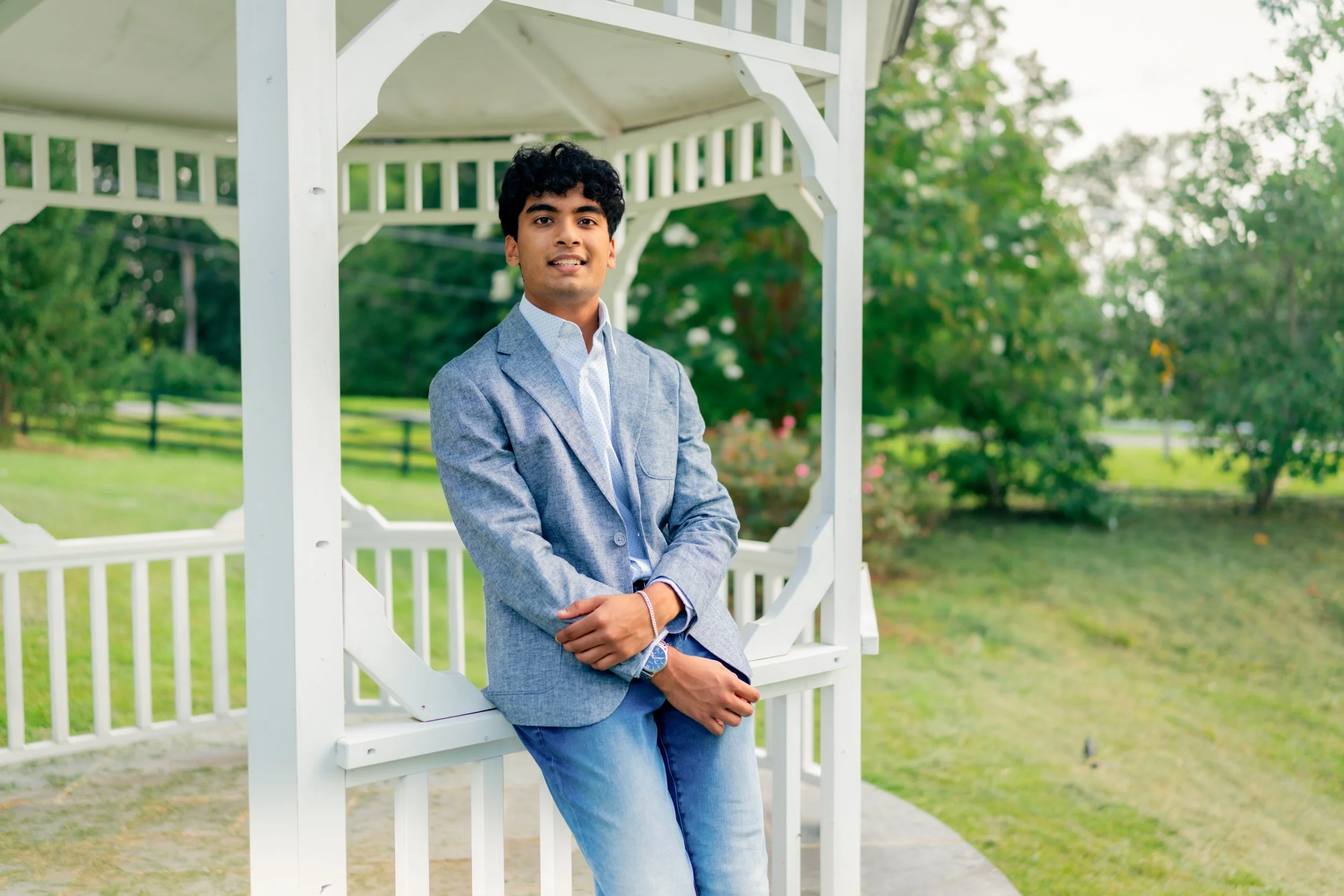 Young man in blue blazer and jeans standing inside a white gazebo in a park with trees in the background.