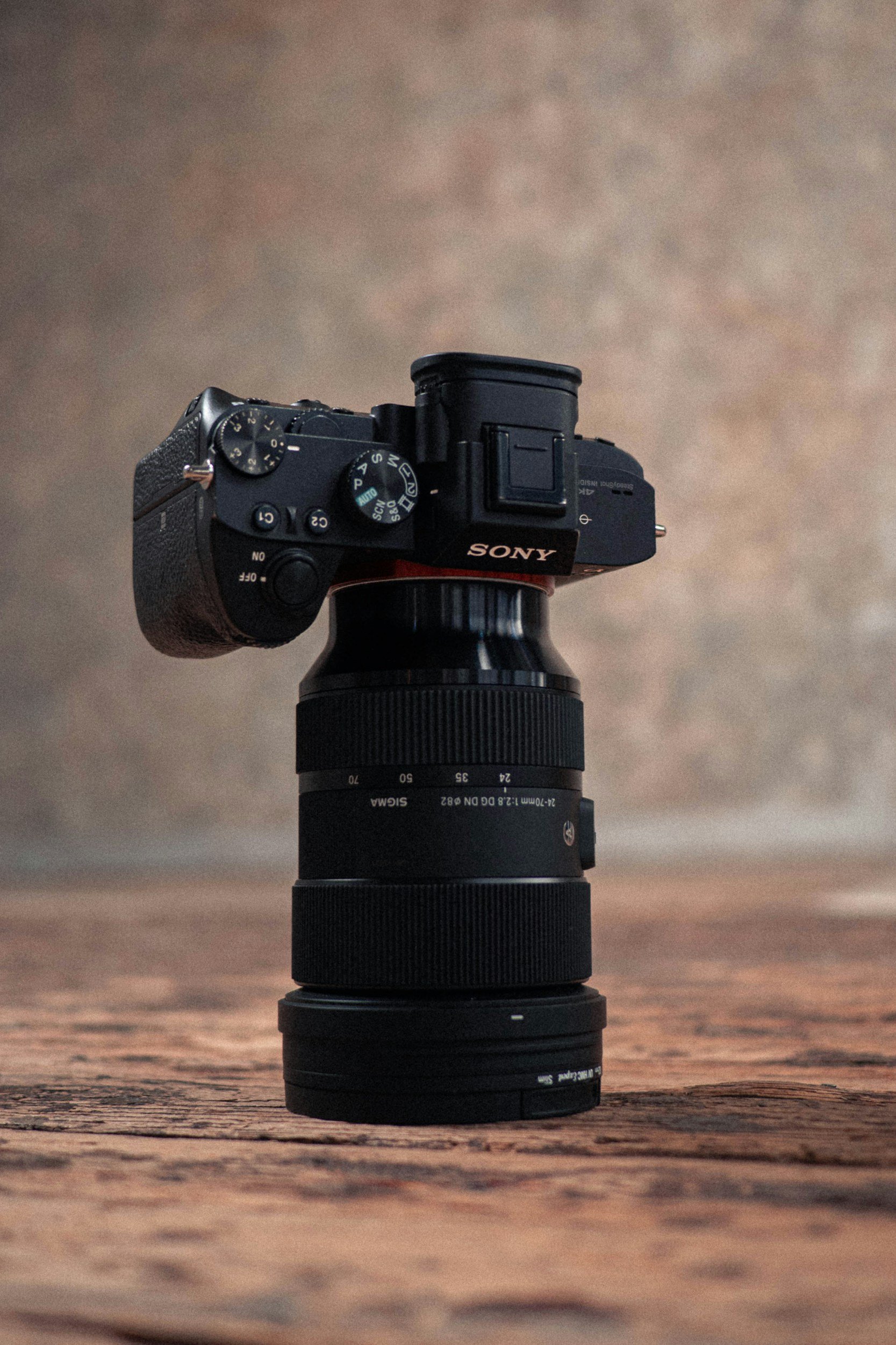 A Sony digital camera mounted on a camera lens, balanced on a stacked set of camera lenses, with a calm body of water and a cloudy sky in the background.