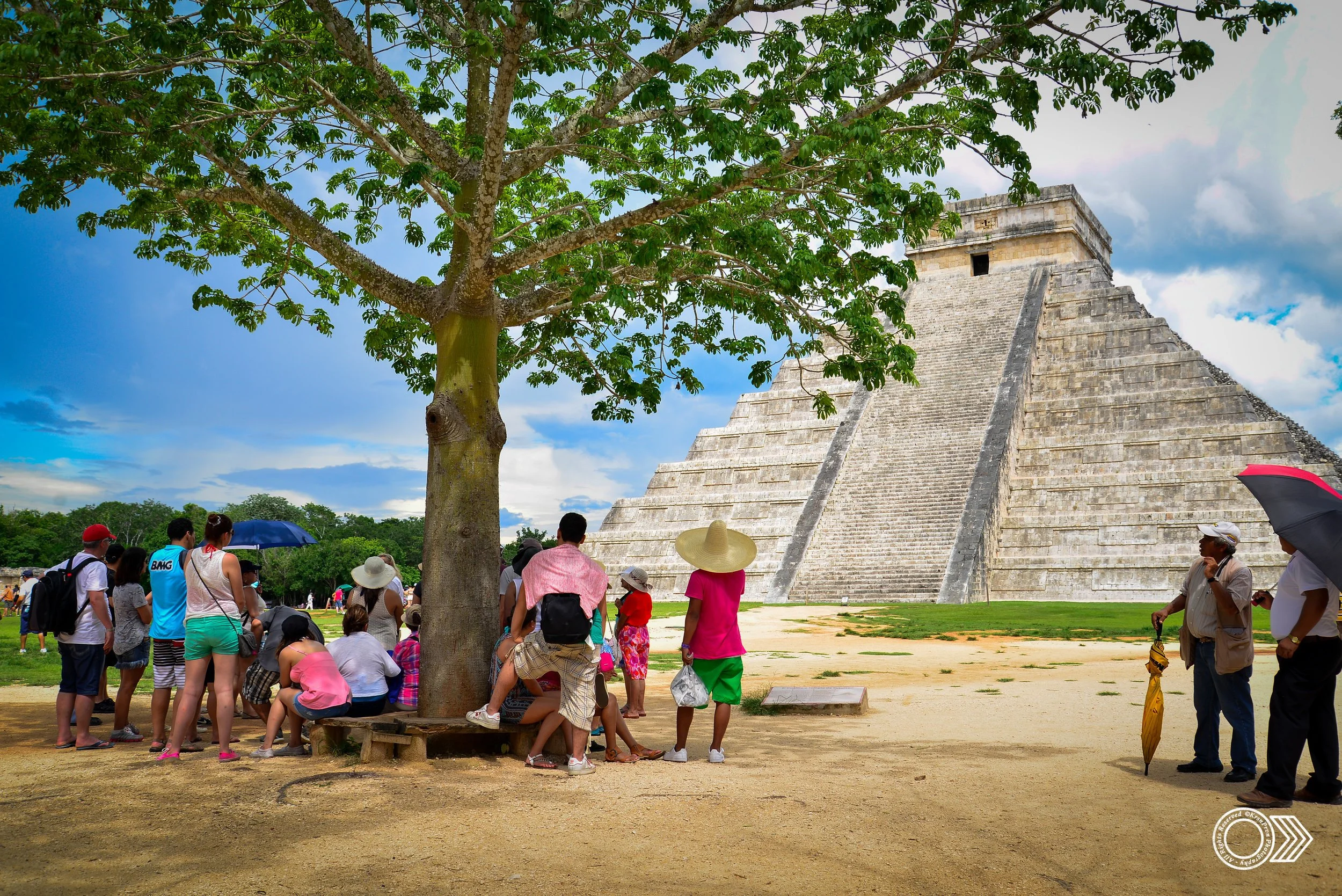 Tourists gathered in front of the ancient Mayan pyramid Chichen Itza in Mexico under a large tree, with some holding umbrellas and wearing hats, against a partly cloudy sky.