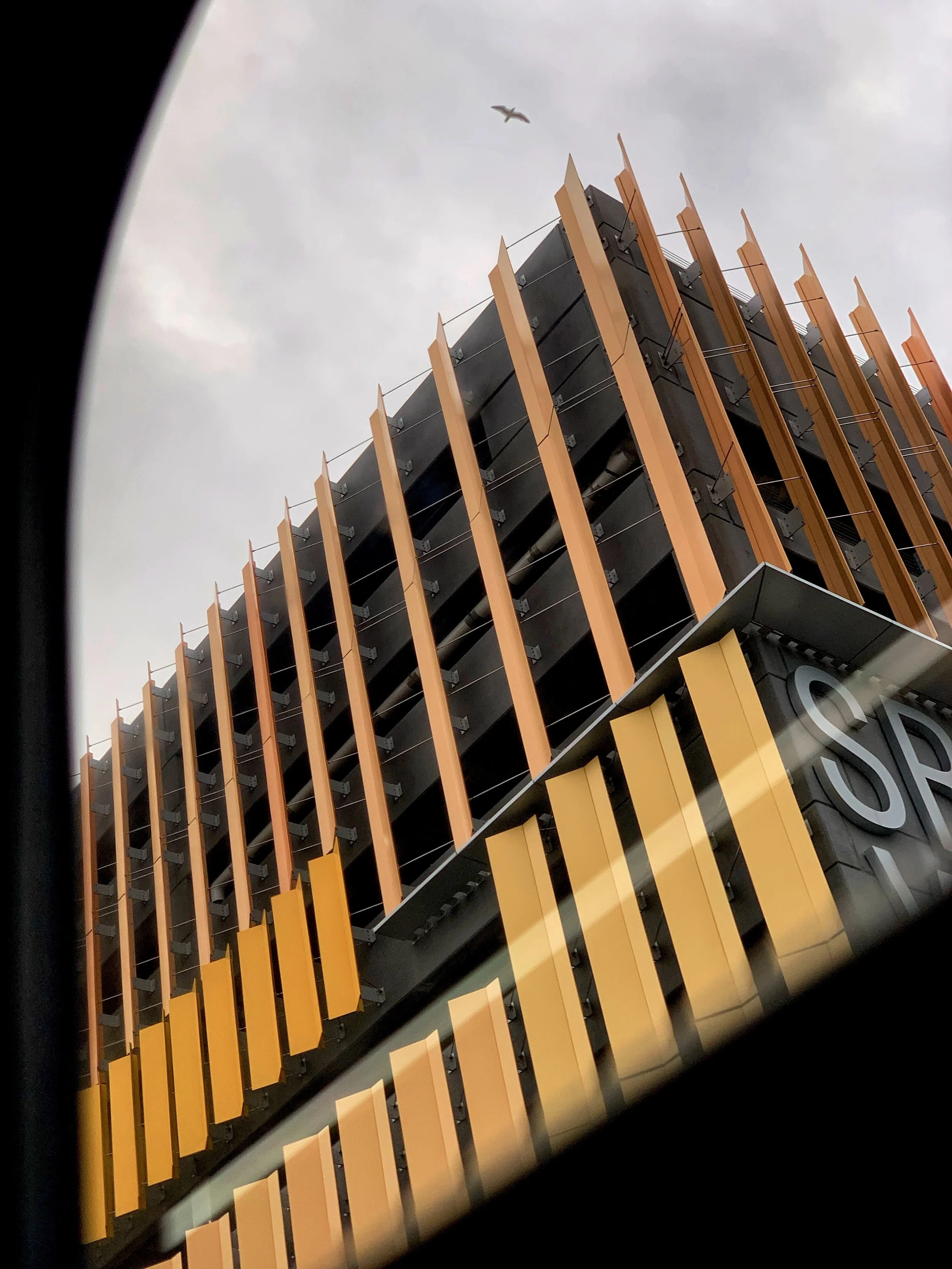 A photo looking up at a modern building with orange vertical fins, gray panels, and a sign that says 'S'. There is a bird flying in the cloudy sky above.