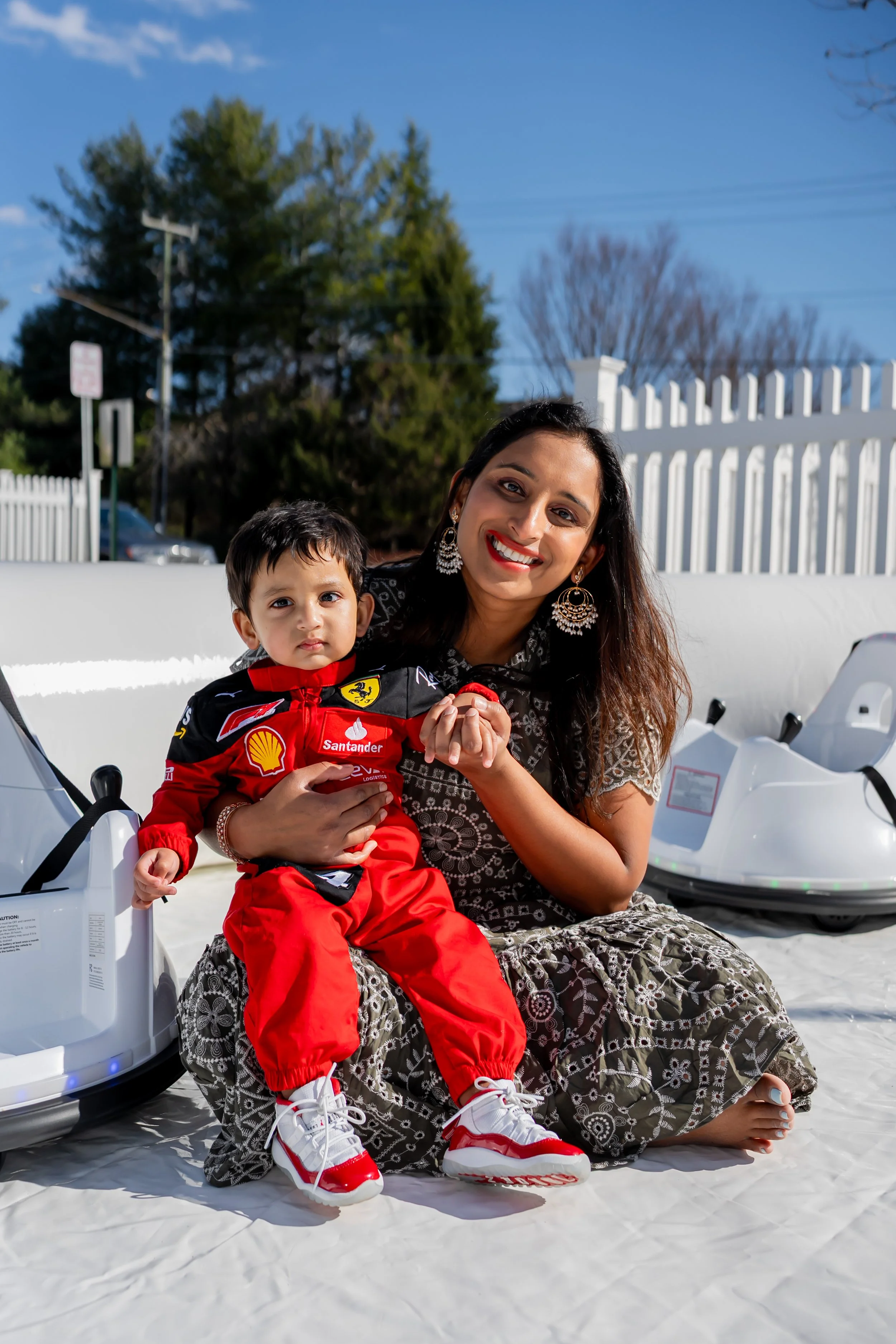 A woman and a young boy sitting on a white surface outdoors, with blue sky, trees, and white fencing in the background. The woman is smiling, wearing earrings, a patterned dress, and has long dark hair. The boy is wearing a red racing suit with logos