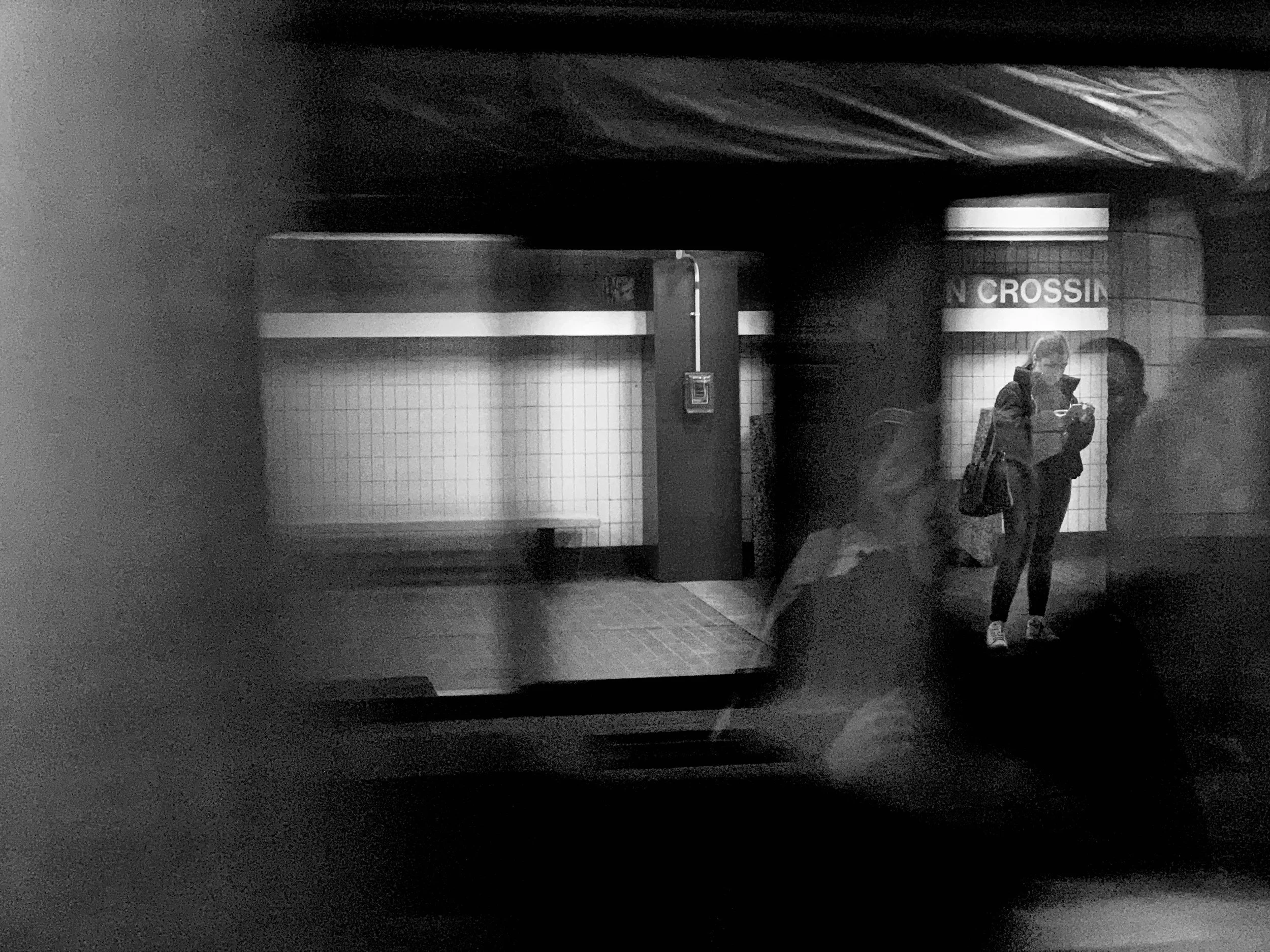 Two people standing inside a subway station, wearing face masks, with tiled walls and a sign that reads "CROSSIN".