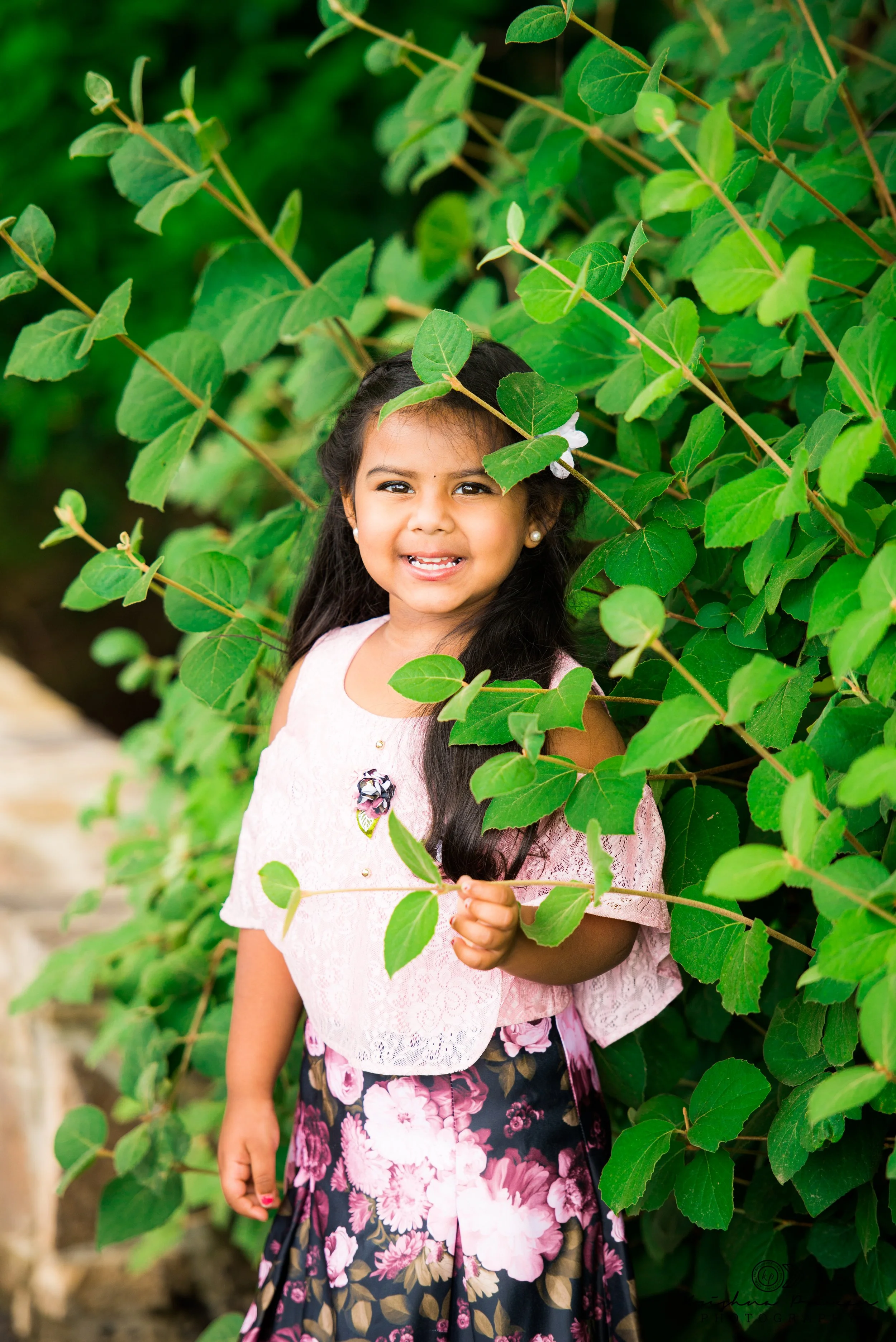 A young girl with long dark hair, wearing a pink lace top and a floral skirt, stands among green leafy bushes, smiling at the camera.