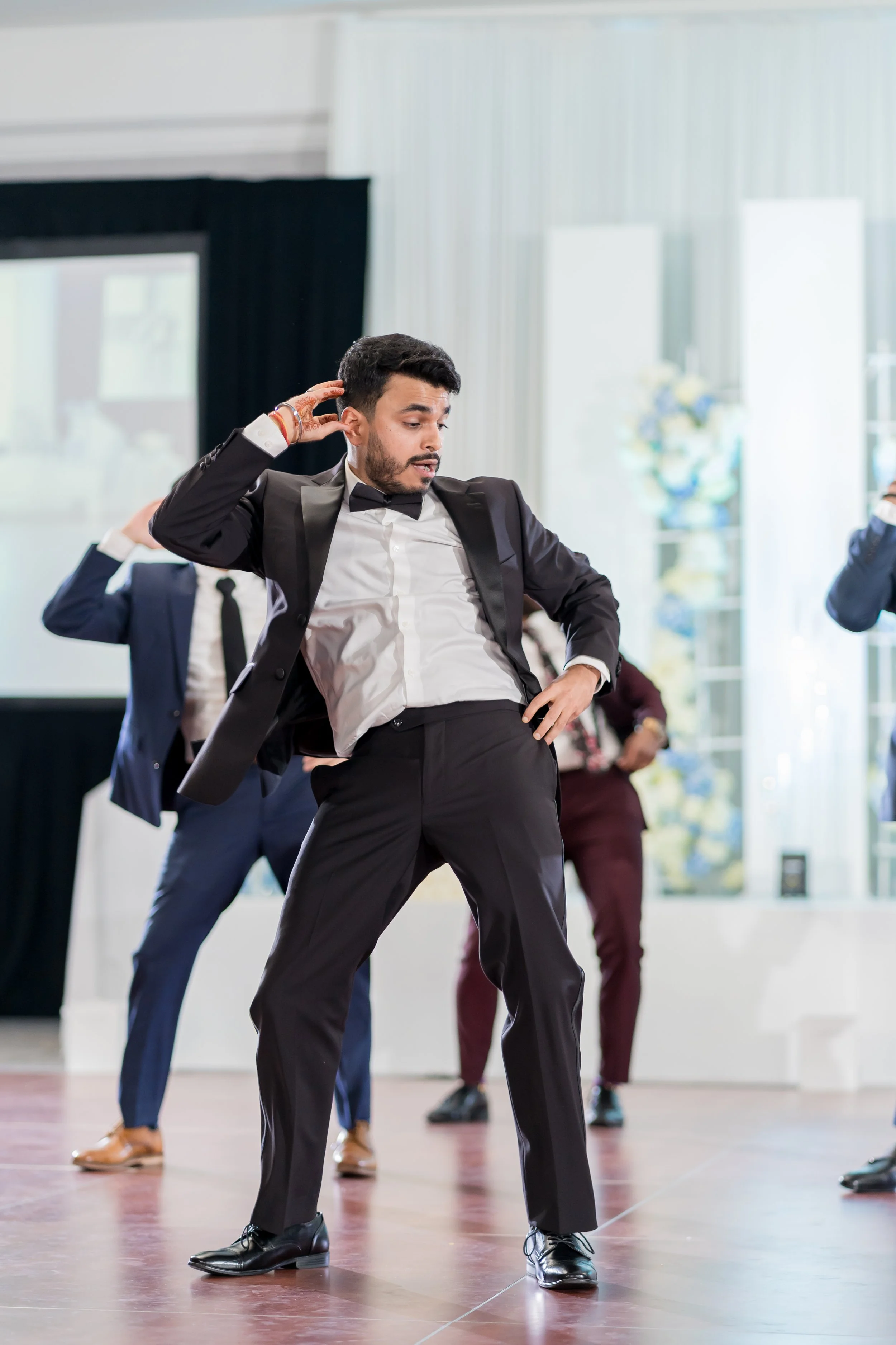 A man dressed in a tuxedo performing a dance move at an indoor event, with other people in formal attire in the background.