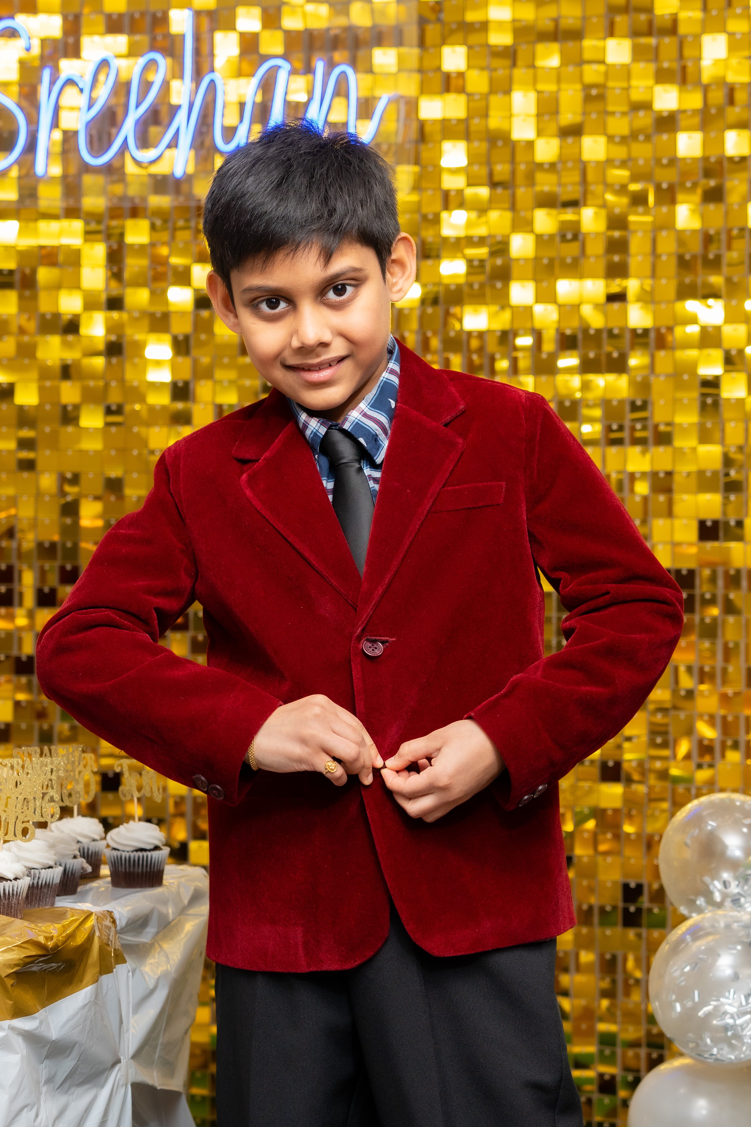 A young boy with dark hair, wearing a red velvet blazer, a blue plaid shirt, and a black tie, standing in front of a gold and yellow background with a neon sign that reads 'spreeen'.