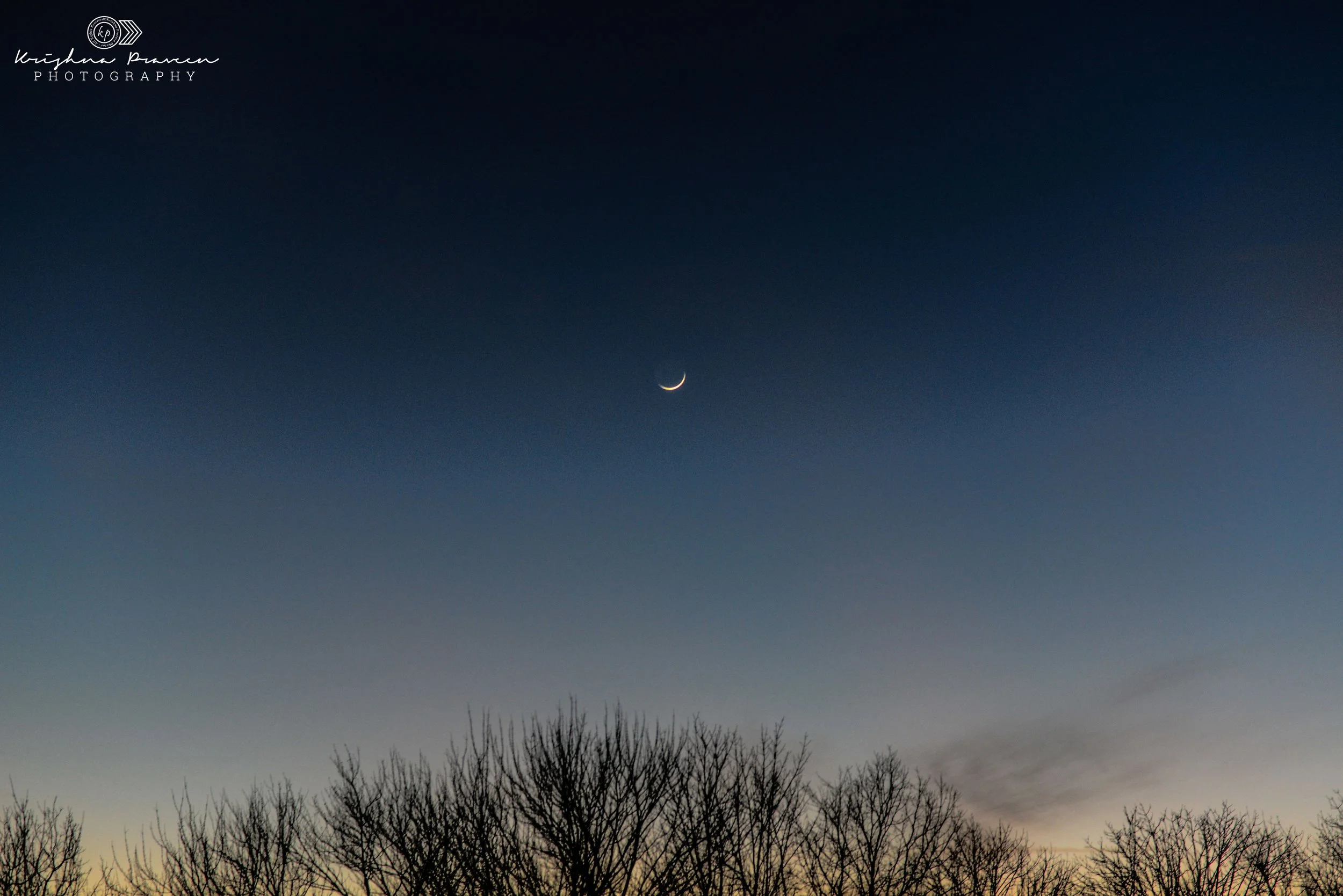 The image depicts a clear night sky with a crescent moon and scattered clouds. In the foreground, there are silhouettes of leafless trees, and the horizon shows a slight yellowish glow, possibly from the setting or rising sun.