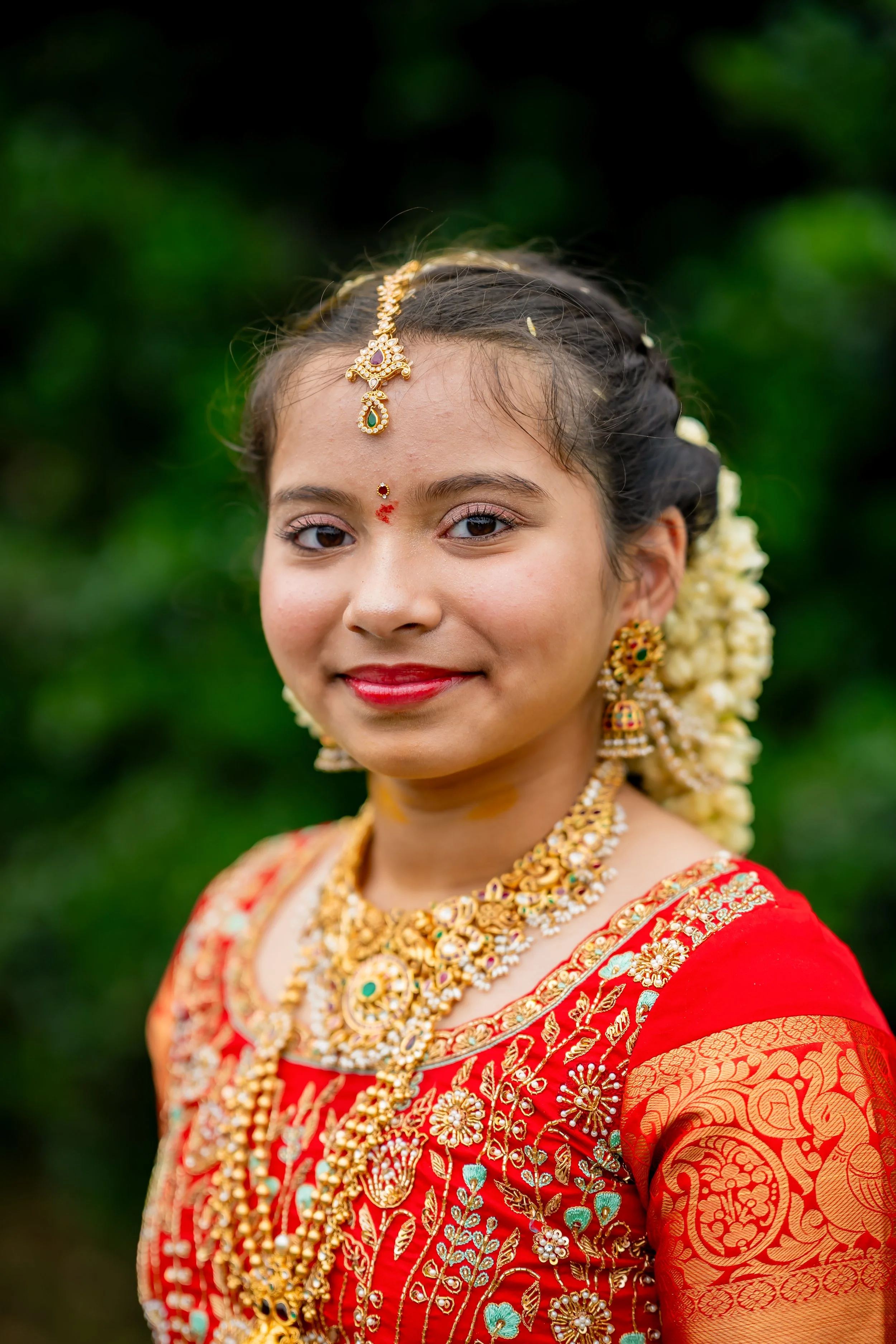 Woman dressed in traditional red and gold Indian wedding attire with intricate jewelry, including necklaces, earrings, and a headpiece, standing outdoors with a blurred green background.