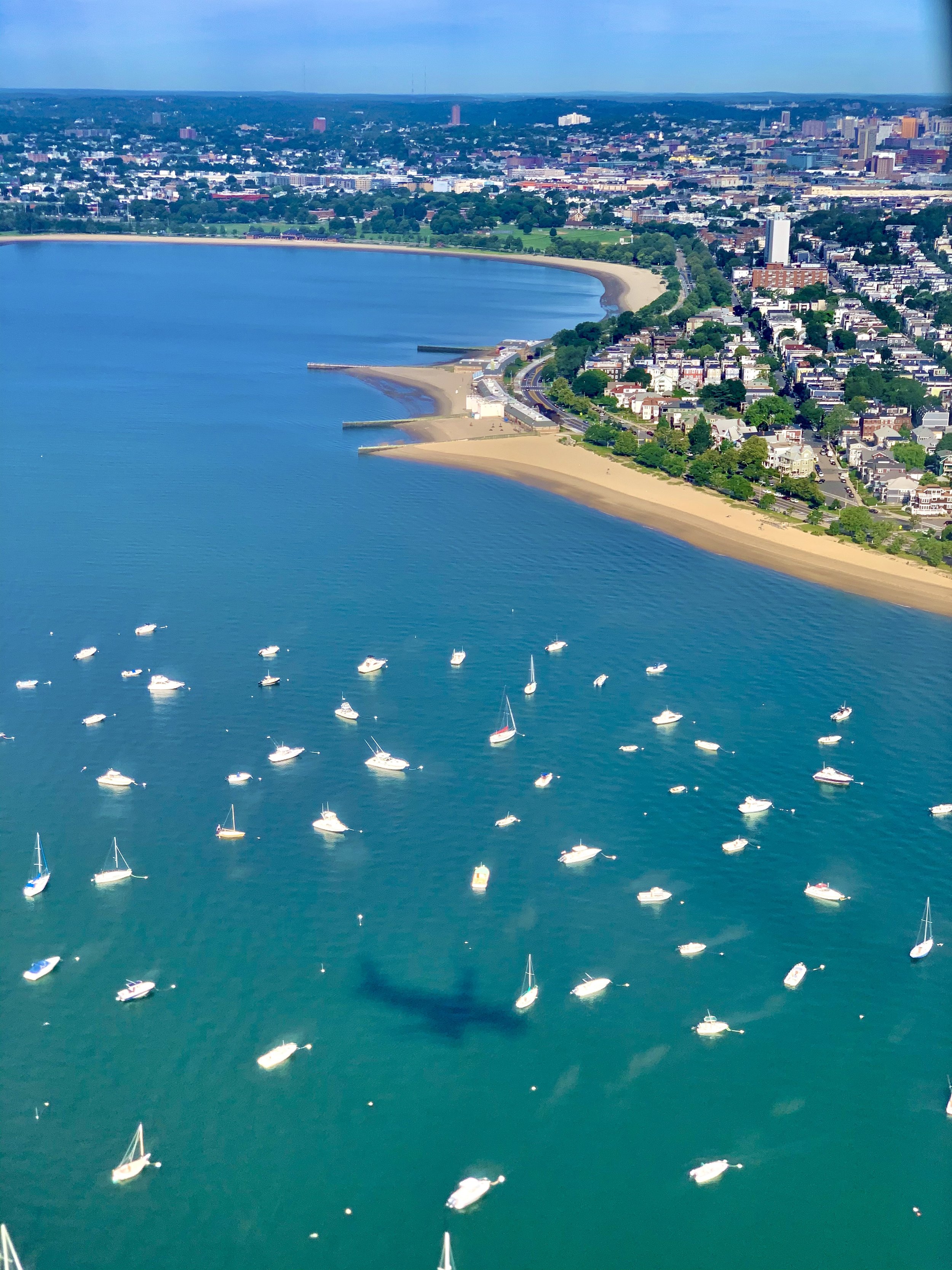 Aerial view of a coastal city with a bay, sandy beach, and numerous sailboats in the water. The city skyline features various buildings and a park area along the shoreline.
