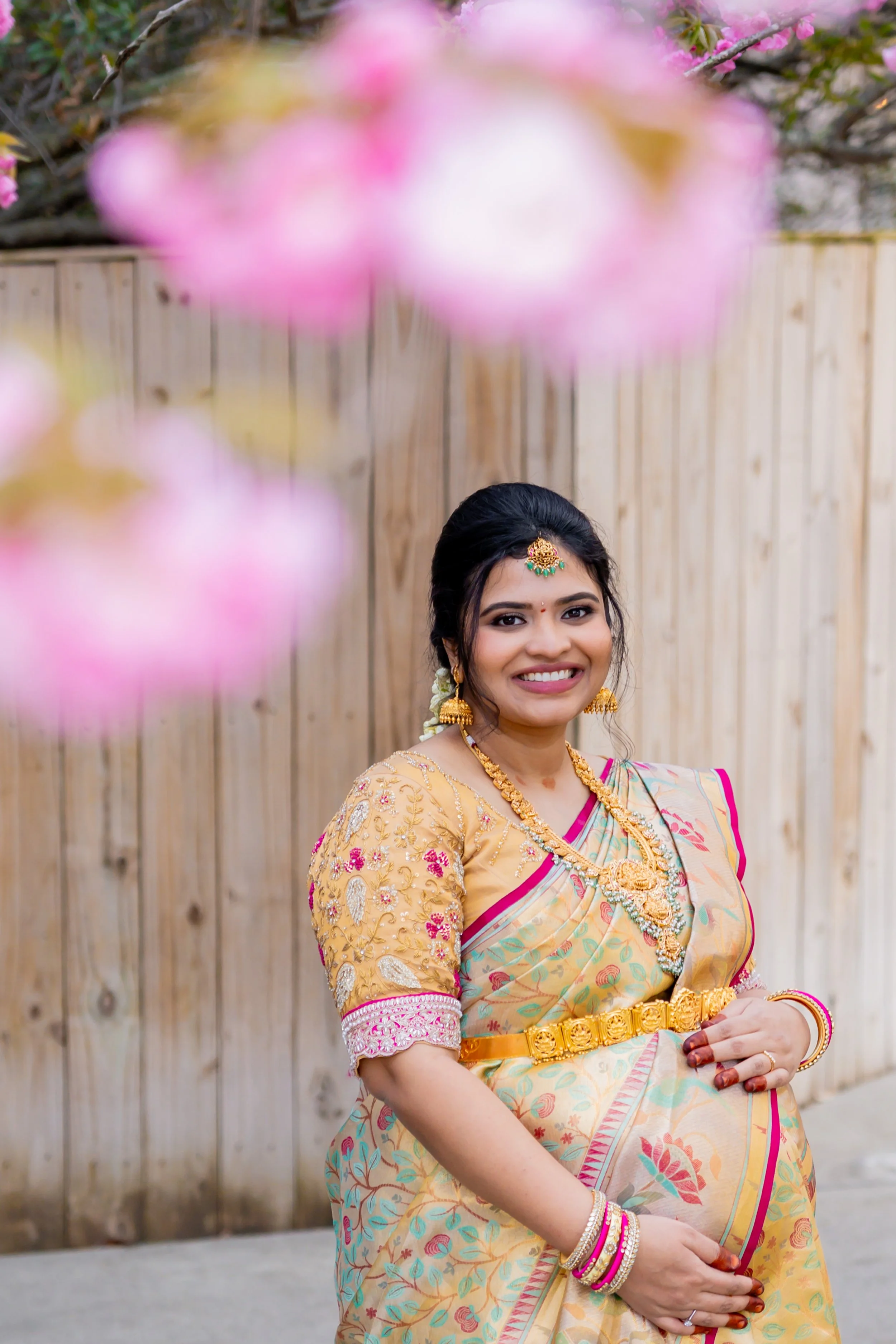 A woman dressed in traditional colorful Indian attire, wearing jewelry, standing outdoors in front of a wooden fence, with pink blossoms in the foreground.