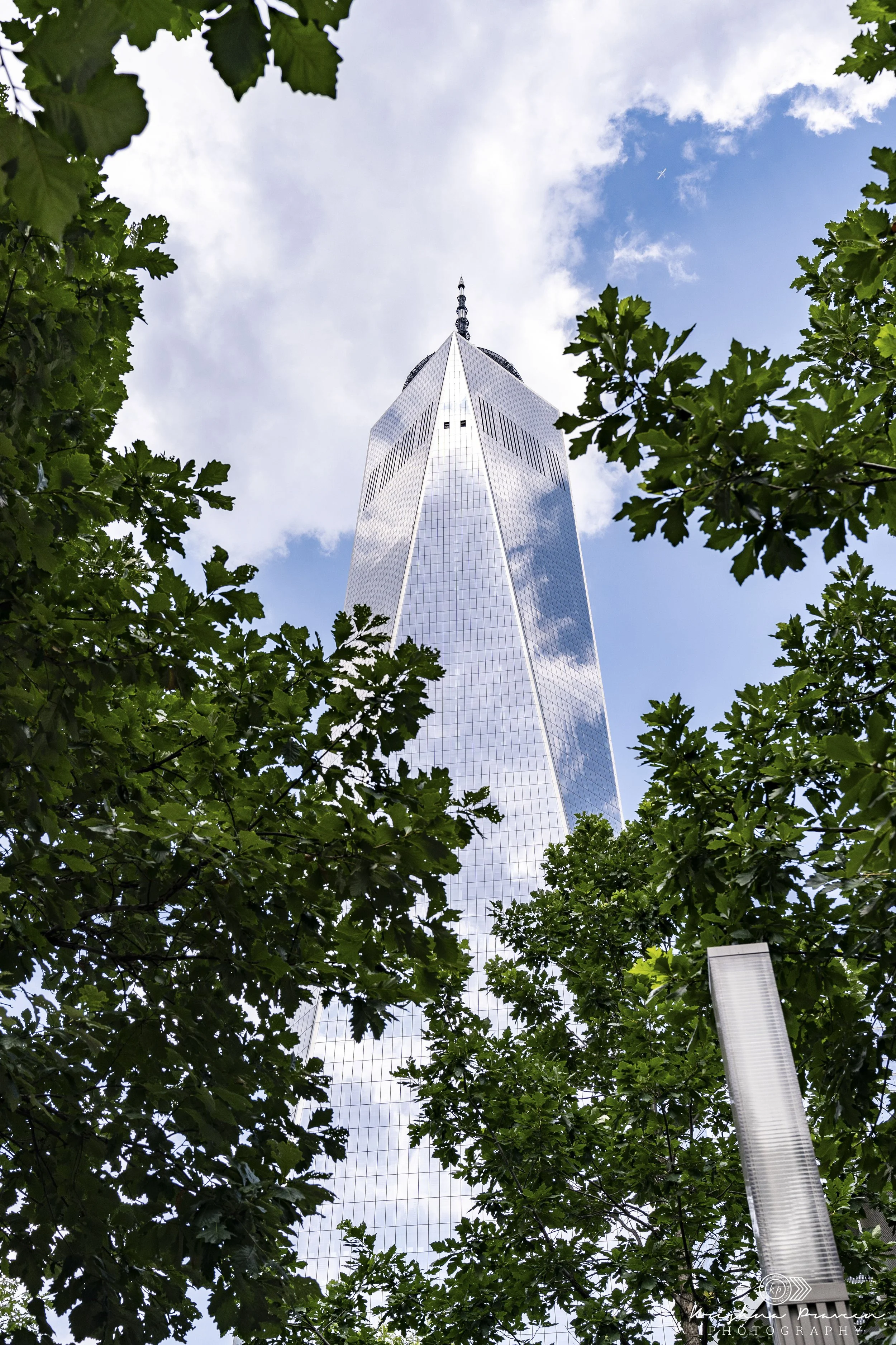 View looking up at One World Trade Center in New York City, framed by green tree leaves and a partially cloudy sky with a small airplane in the distance.