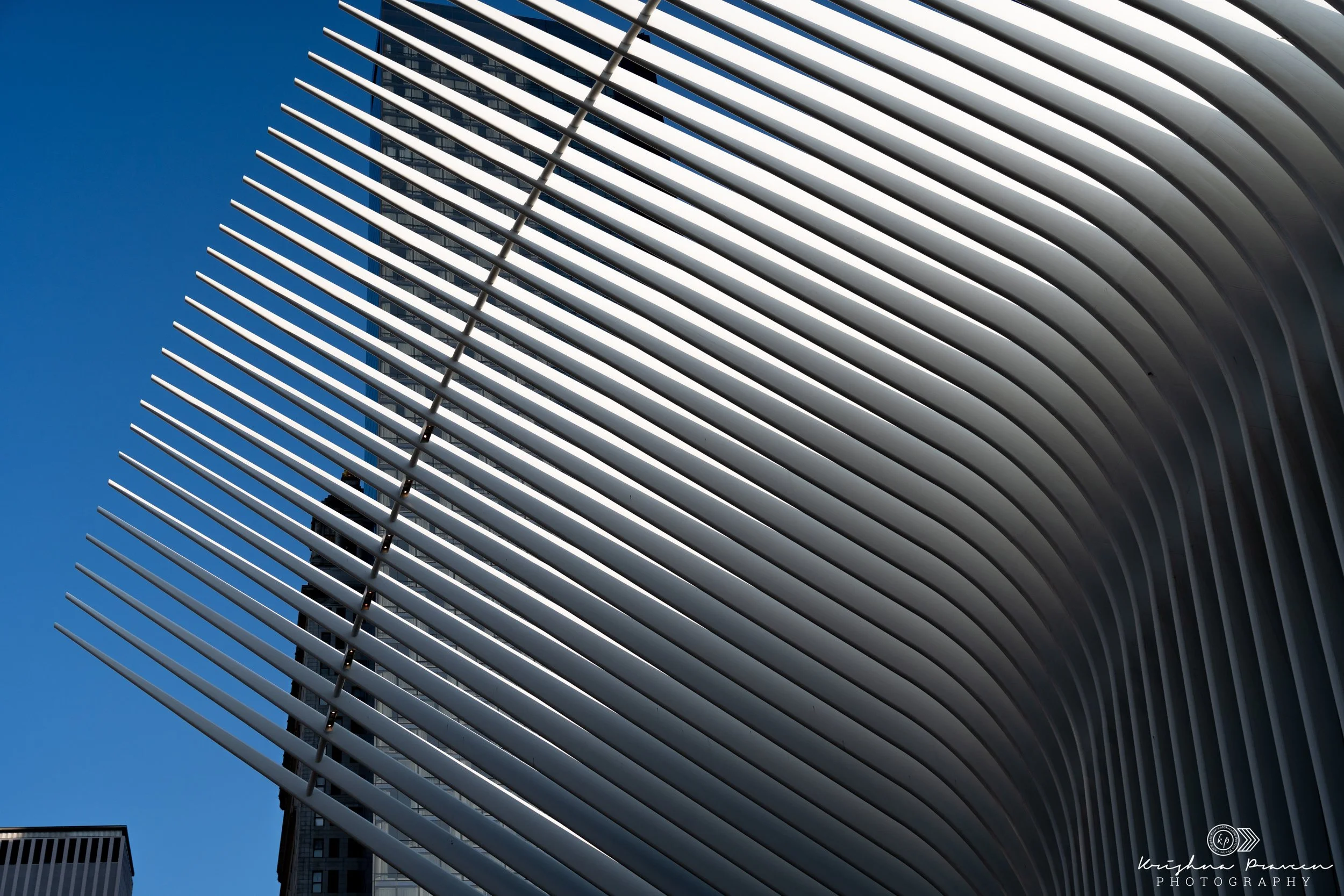 Modern architectural building with white horizontal slats or louvers against a clear blue sky, with some taller buildings partially visible in the background.