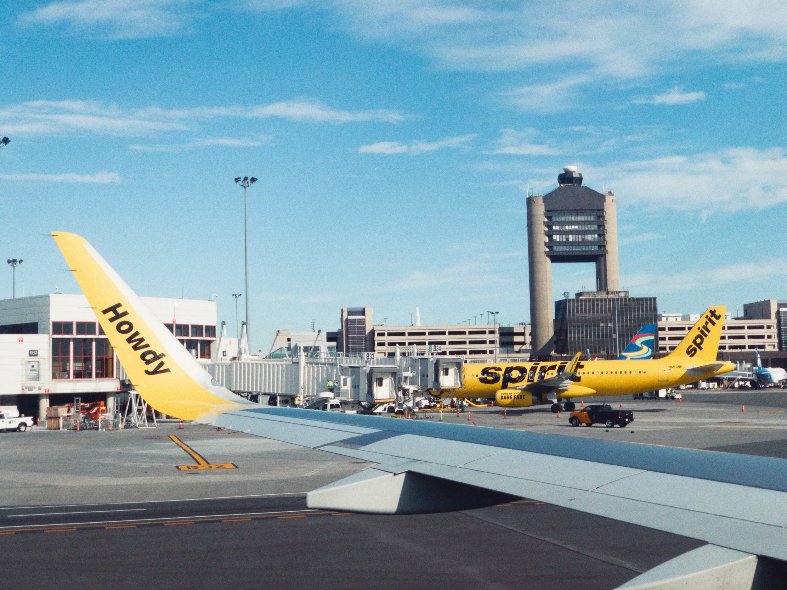 View from airplane window showing airport tarmac with yellow Spirit Airlines plane, airport buildings, and a control tower under a partly cloudy sky.