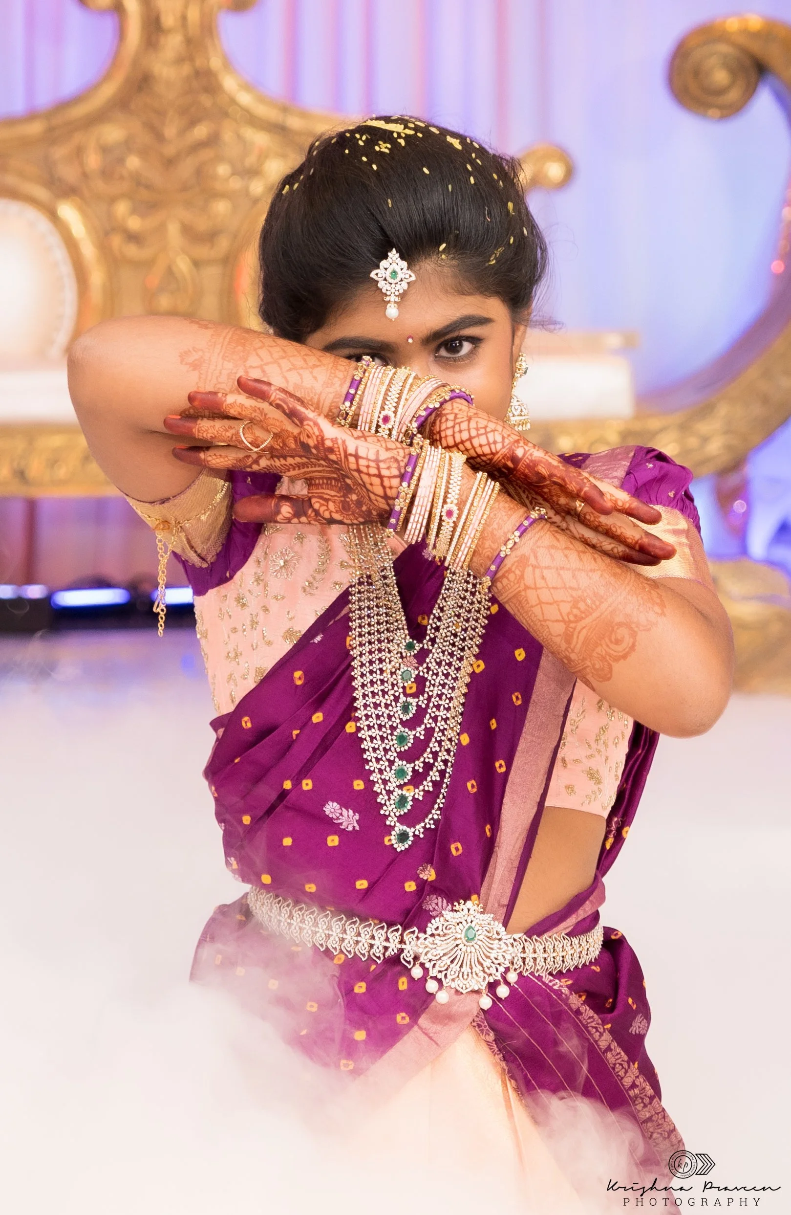A young woman wearing traditional Indian attire, a purple and gold saree, with intricate jewelry, including earrings, a headpiece, layered necklaces, and bangles. She is posed with her arms crossed in front of her face, displaying detailed henna desi