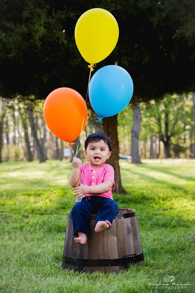 Young child sitting on a wooden barrel outdoors, holding three colorful balloons (yellow, blue, orange), in a park or garden with trees and green grass.