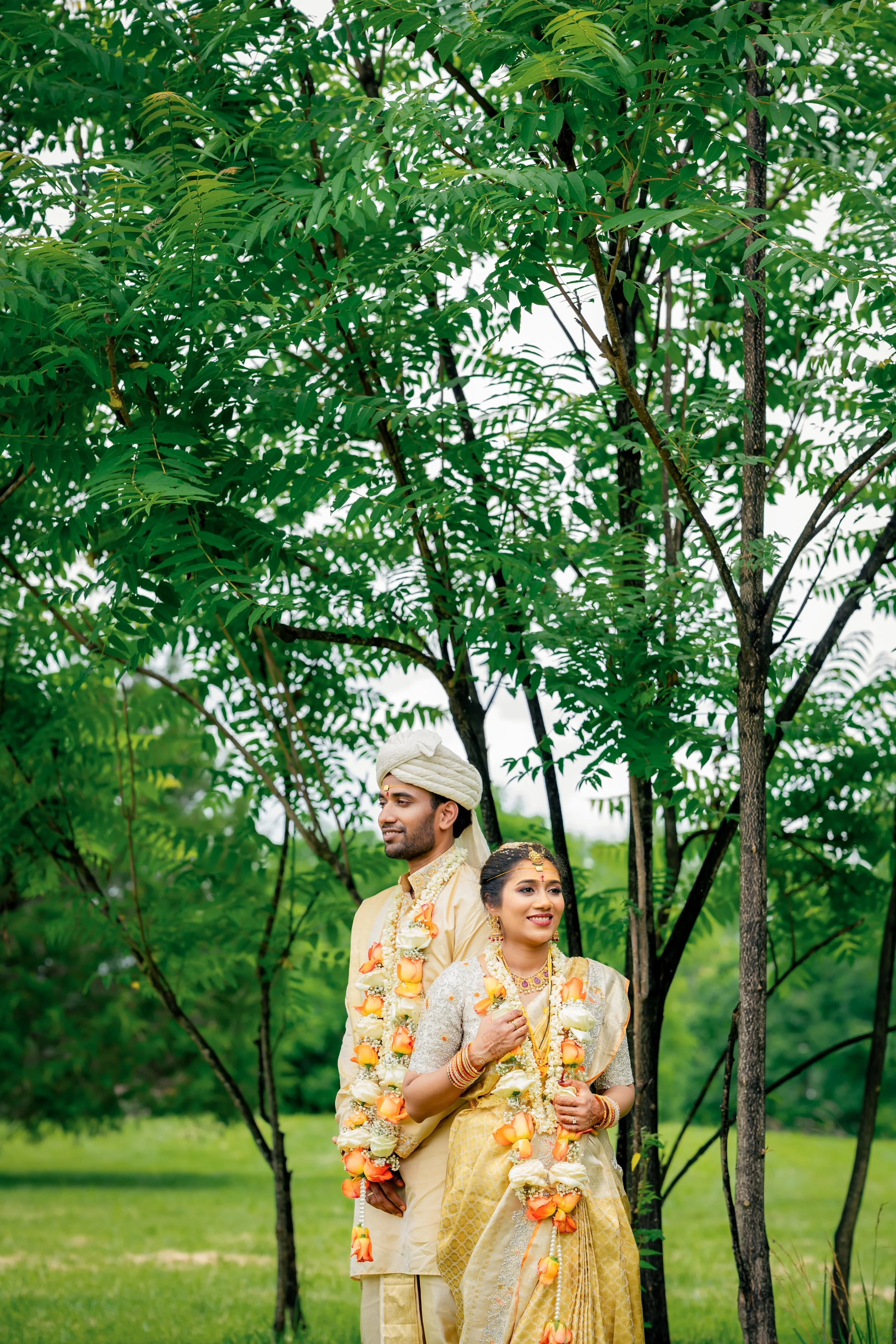 A couple dressed in traditional Indian wedding attire, standing outdoors under a large green tree. They are decorated with flower garlands and jewelry, smiling and looking happy.