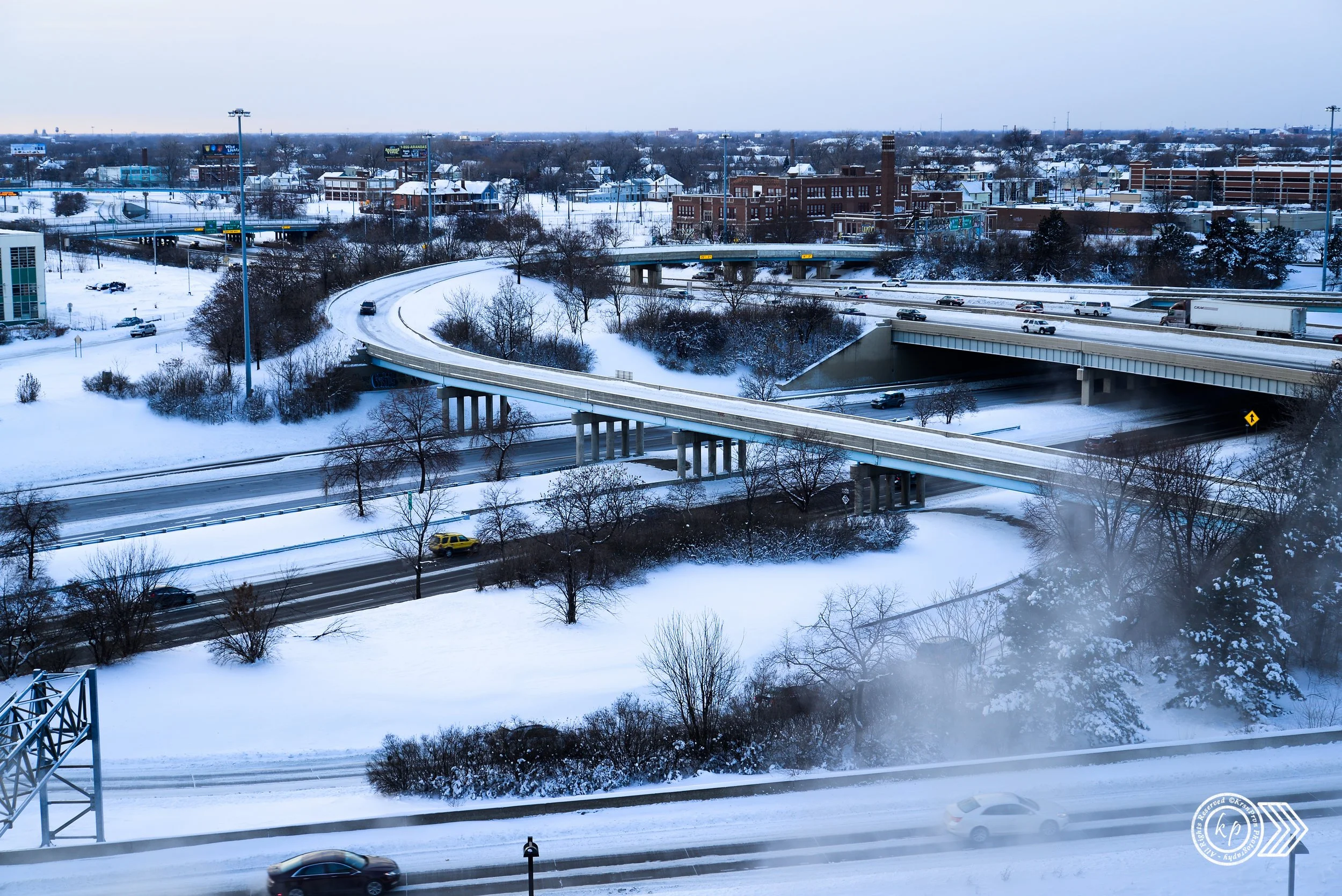 A city highway overpass with multiple lanes, cars driving, and snow-covered roads and trees in the background on a cloudy winter day.