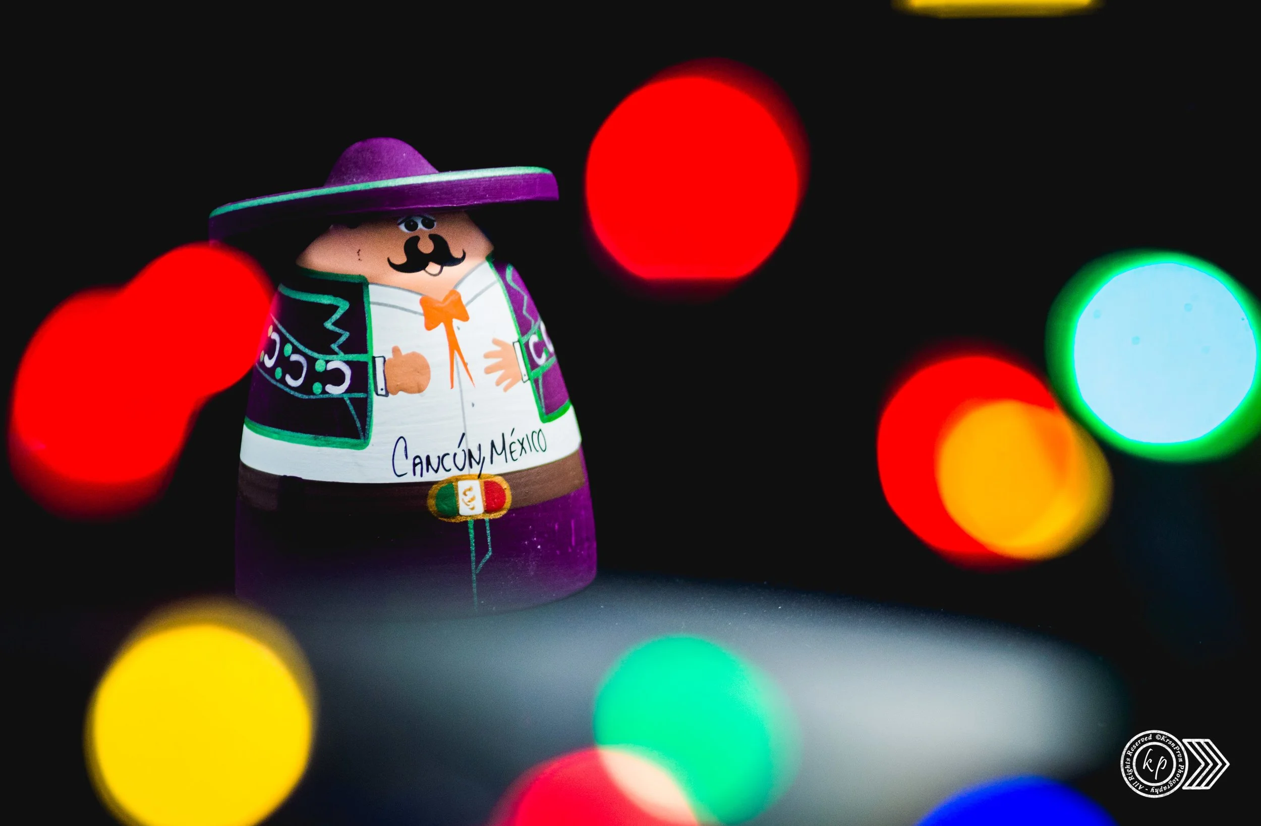Colorful toy figure of a man wearing a purple sombrero and a traditional Mexican outfit with the word 'Cancún, México' written on it, set against a background of blurred multicolored lights.