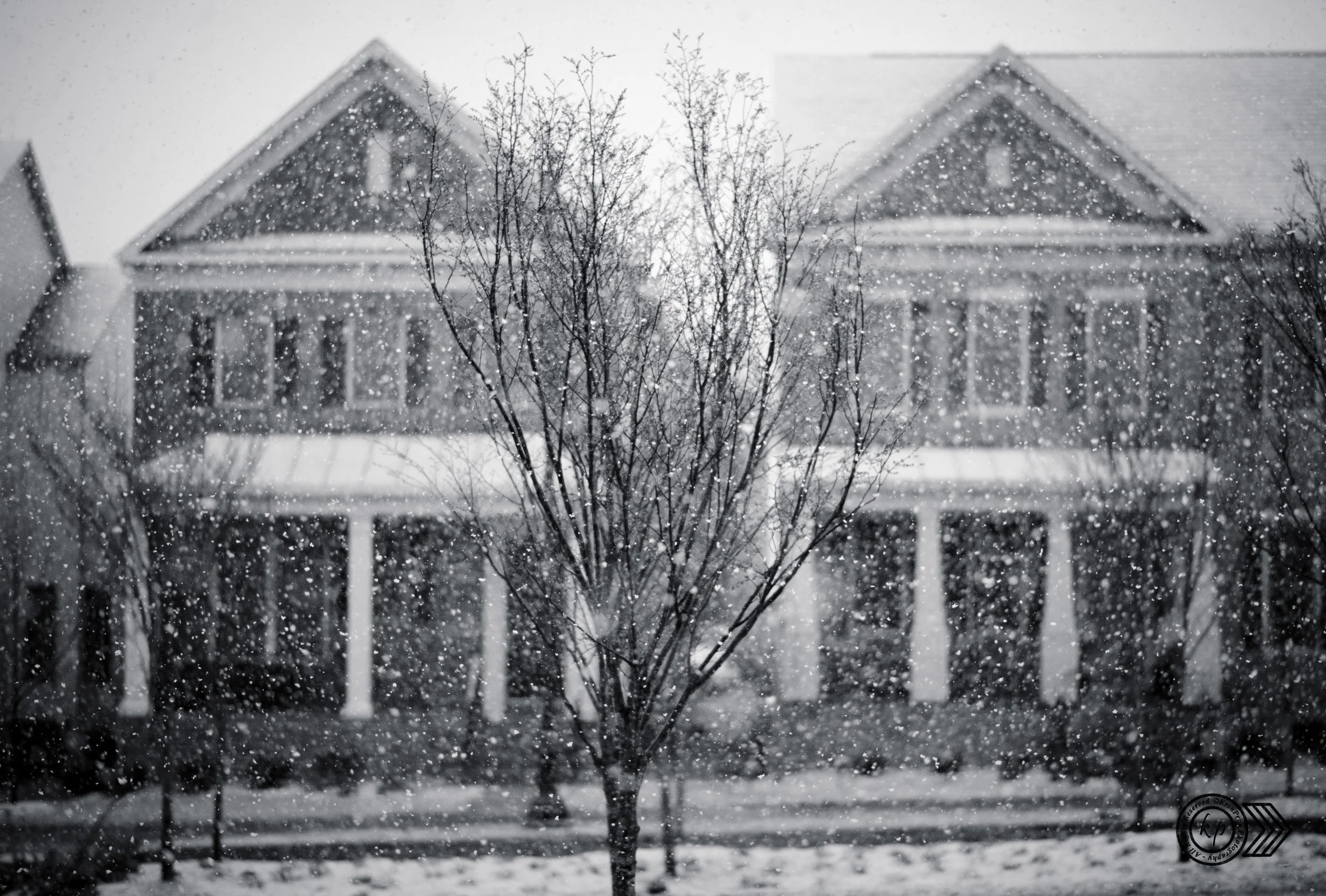 Black and white photograph of a snow-covered residential house with a front porch and porch columns, partially obscured by snow falling and tree branches in the foreground.