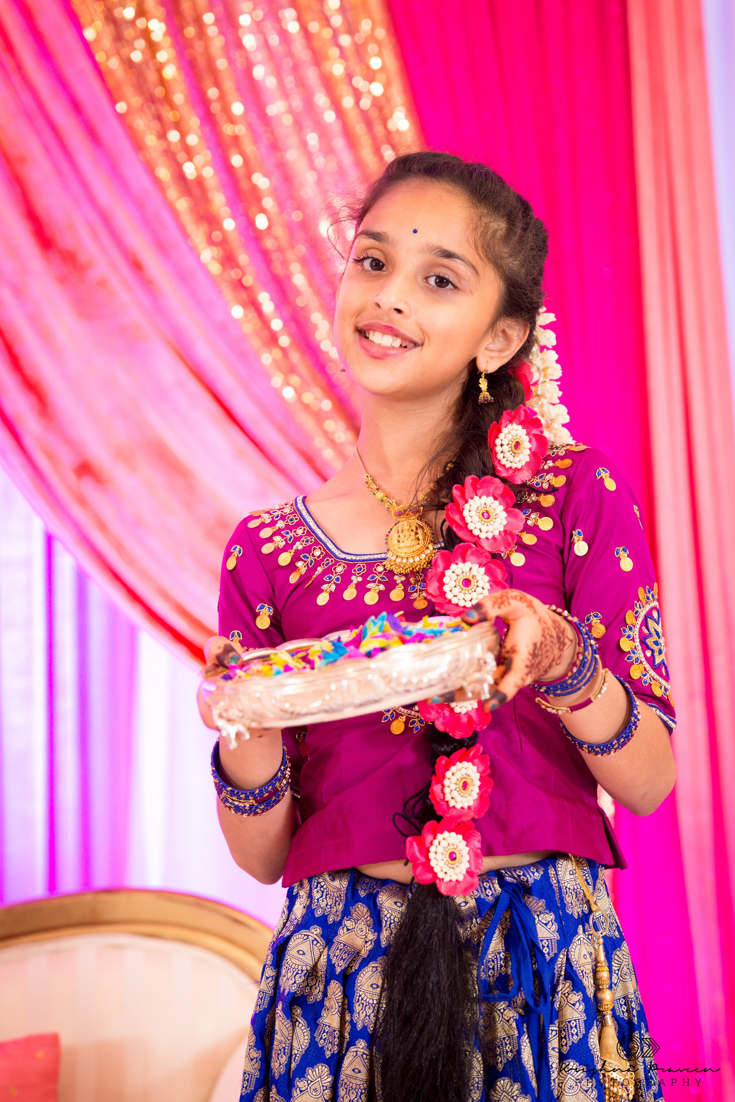 Young girl dressed in traditional Indian attire, smiling, holding a silver plate with colorful items, with a pink and glittery background, decorated with flowers in her hair.