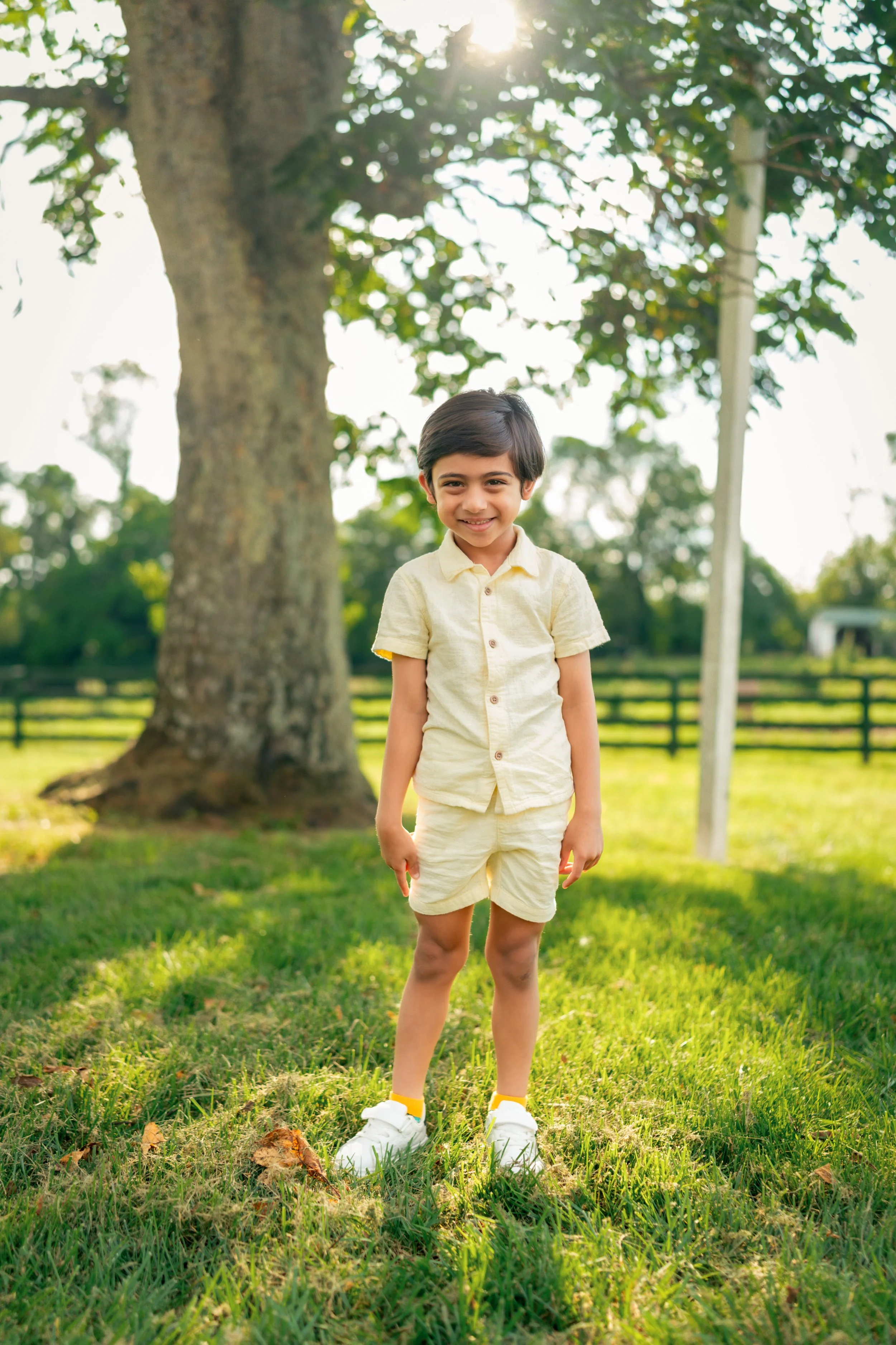 A young boy with dark hair, smiling, wearing a light yellow shirt and shorts, standing on green grass near a large tree under the sunlight.
