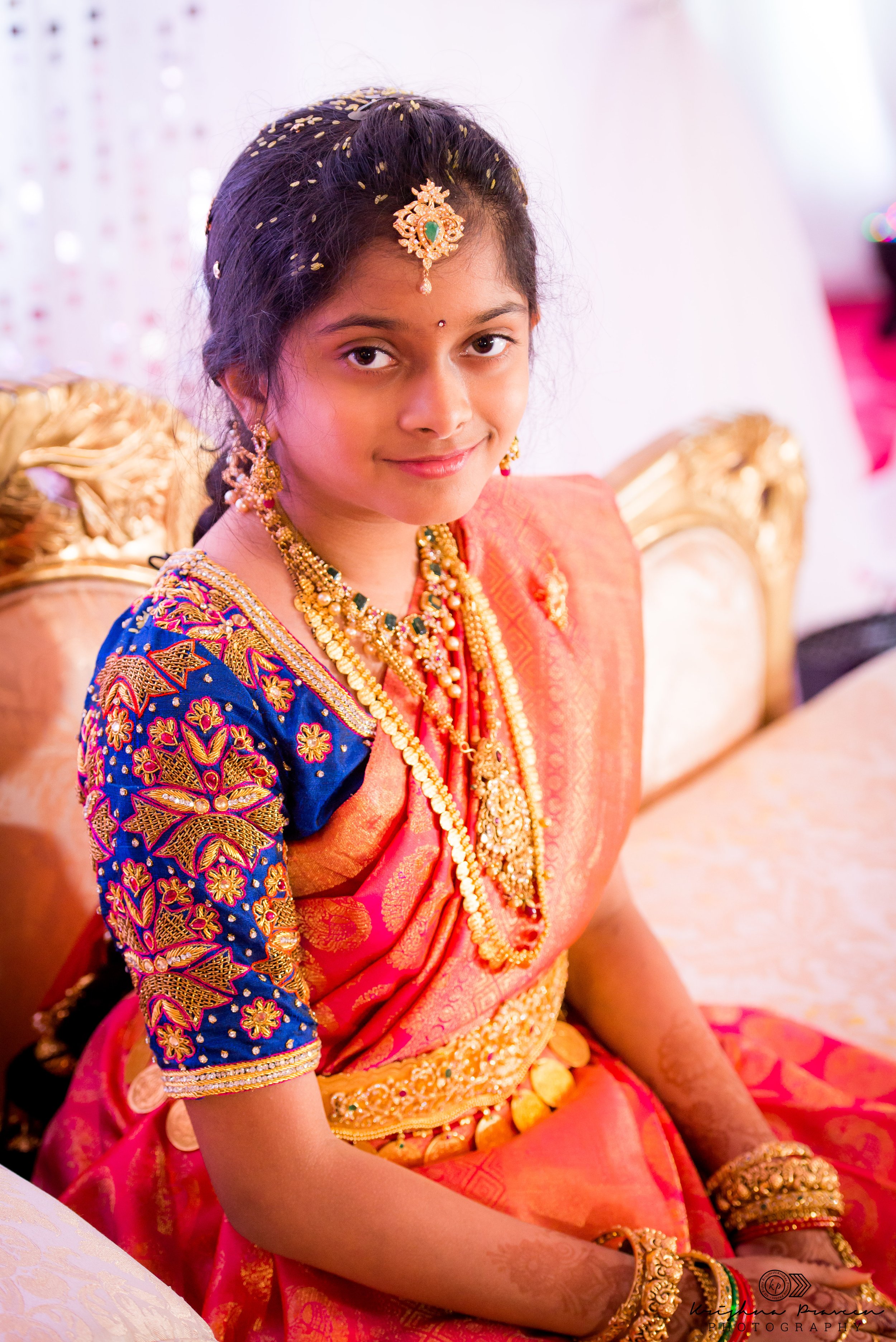 A young girl dressed in traditional Indian attire, wearing vibrant pink and blue embroidered clothes with gold jewelry, sitting on a decorated chair at what appears to be a celebration or wedding.