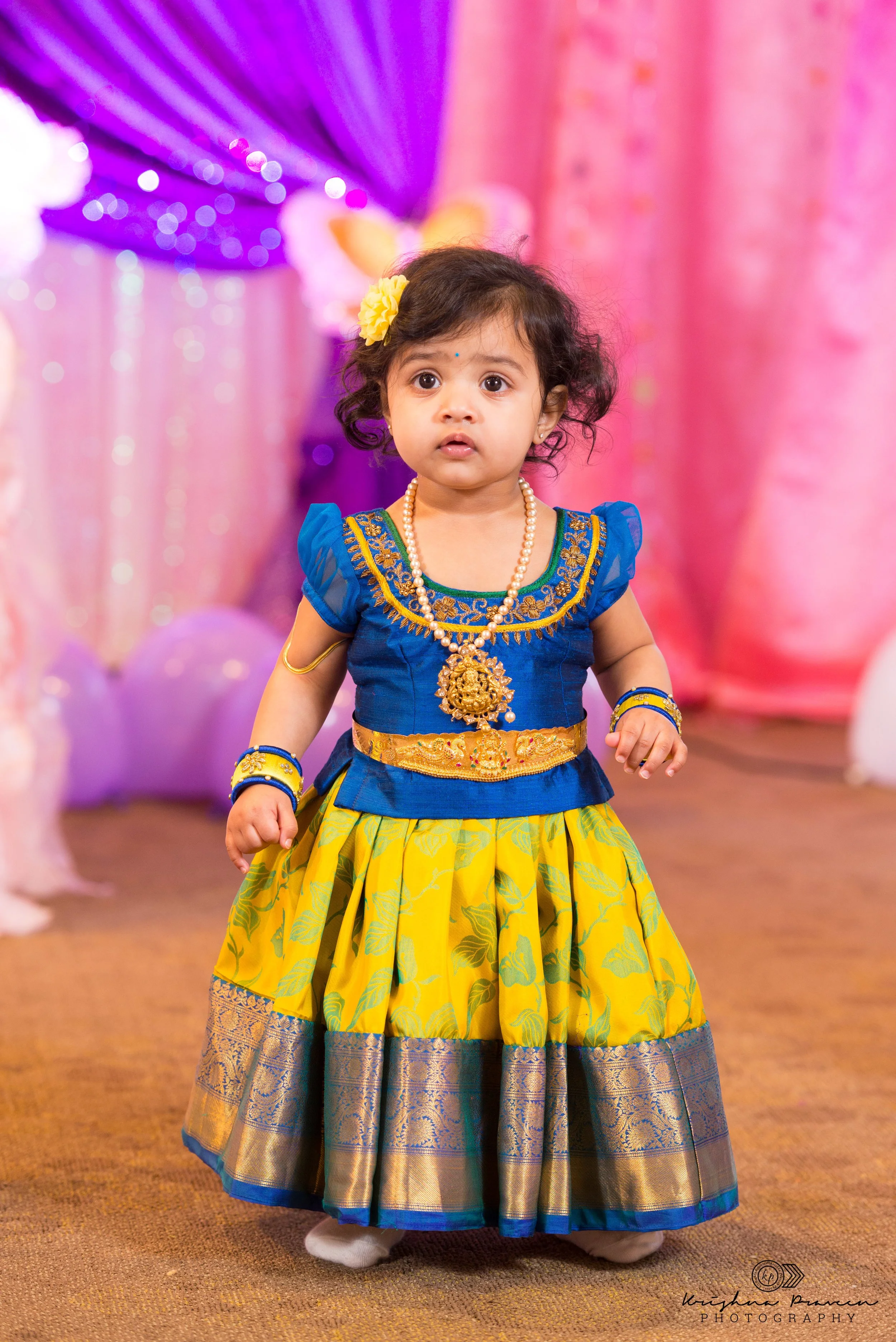A young girl dressed in traditional Indian attire with a blue and yellow skirt, blue top with gold embroidery, and jewelry, standing indoors against pink and purple backdrop decorations.