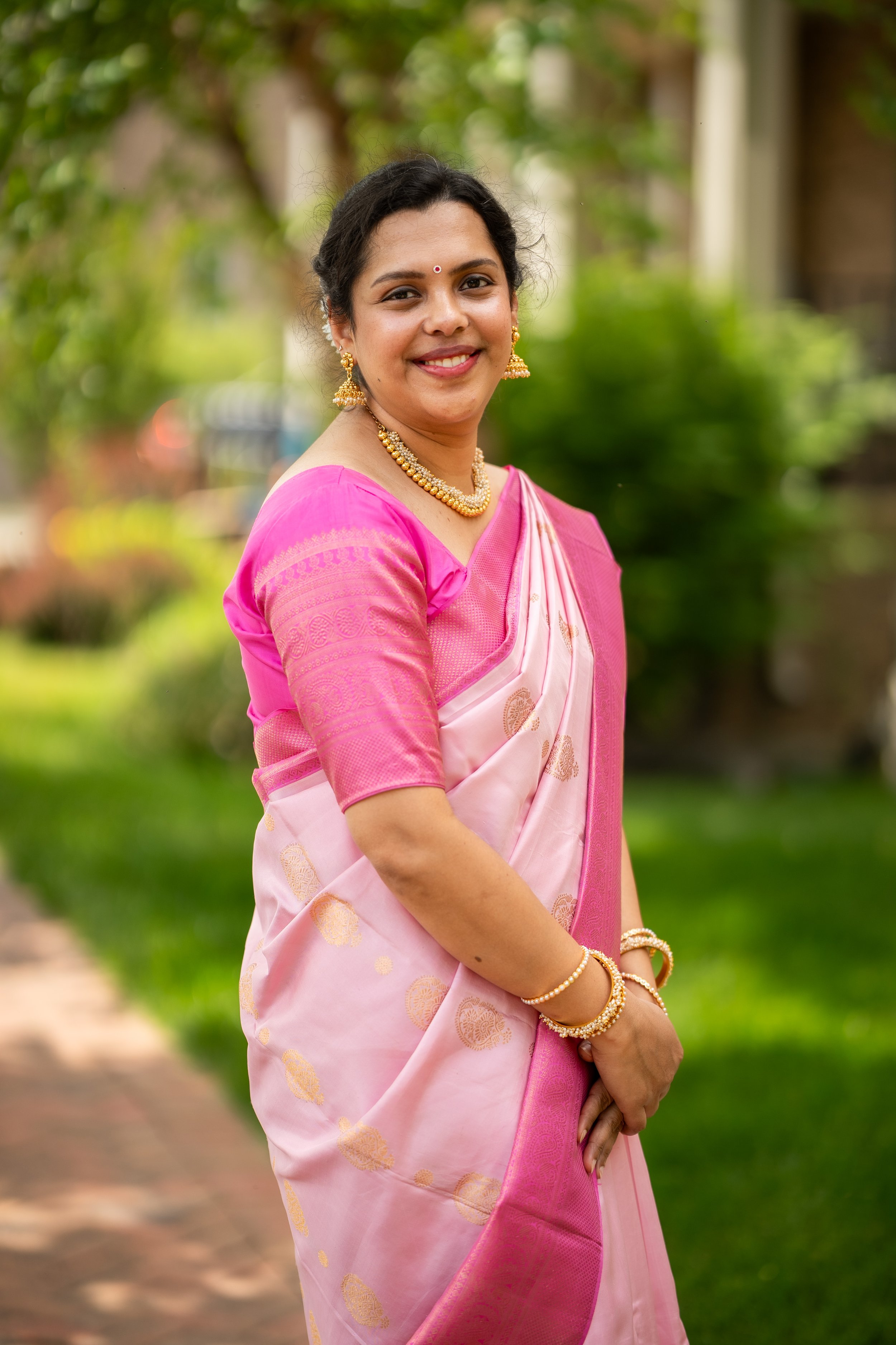 A woman dressed in a pink traditional Indian saree with gold jewelry, smiling outdoors with blurred greenery in the background.