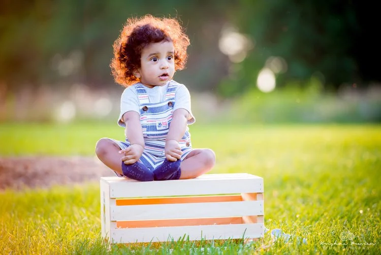 A young child with curly hair sitting cross-legged on a white wooden crate outdoors in a grassy area, with a blurred background of trees and sunlight.