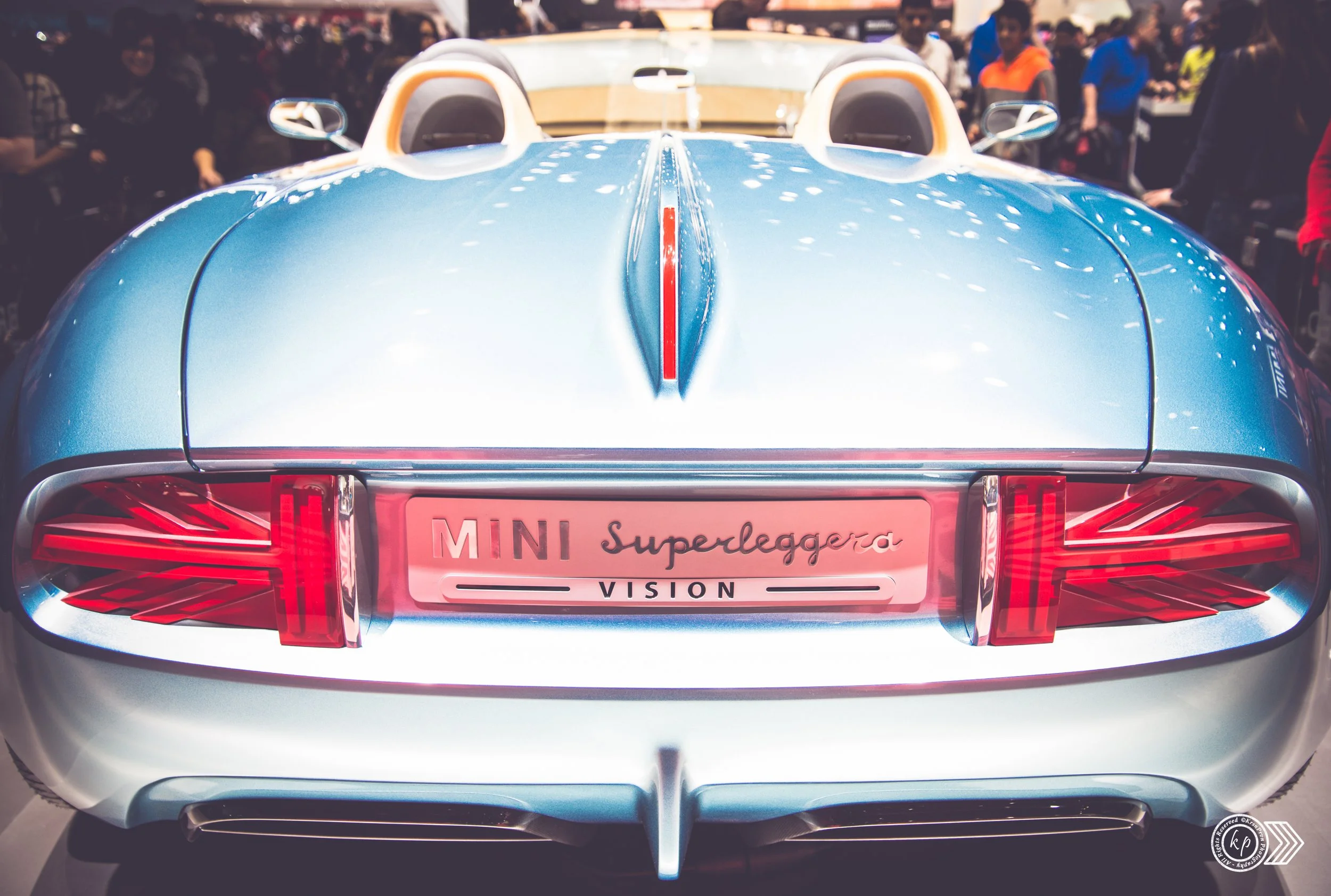 Rear view of a blue sports car with a sleek design at an indoor event, surrounded by a crowd of people.