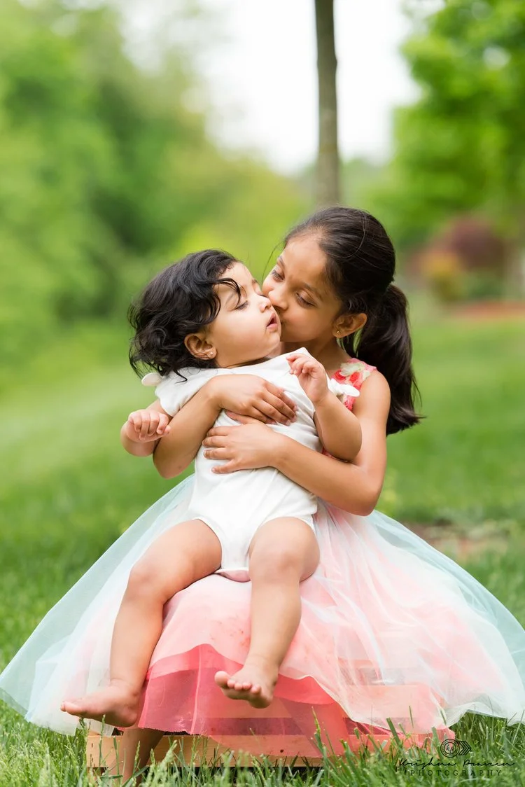 A young girl, dressed in a pastel-colored dress, is hugging and kissing a toddler girl sitting on a wooden box outdoors with green trees and grass in the background.