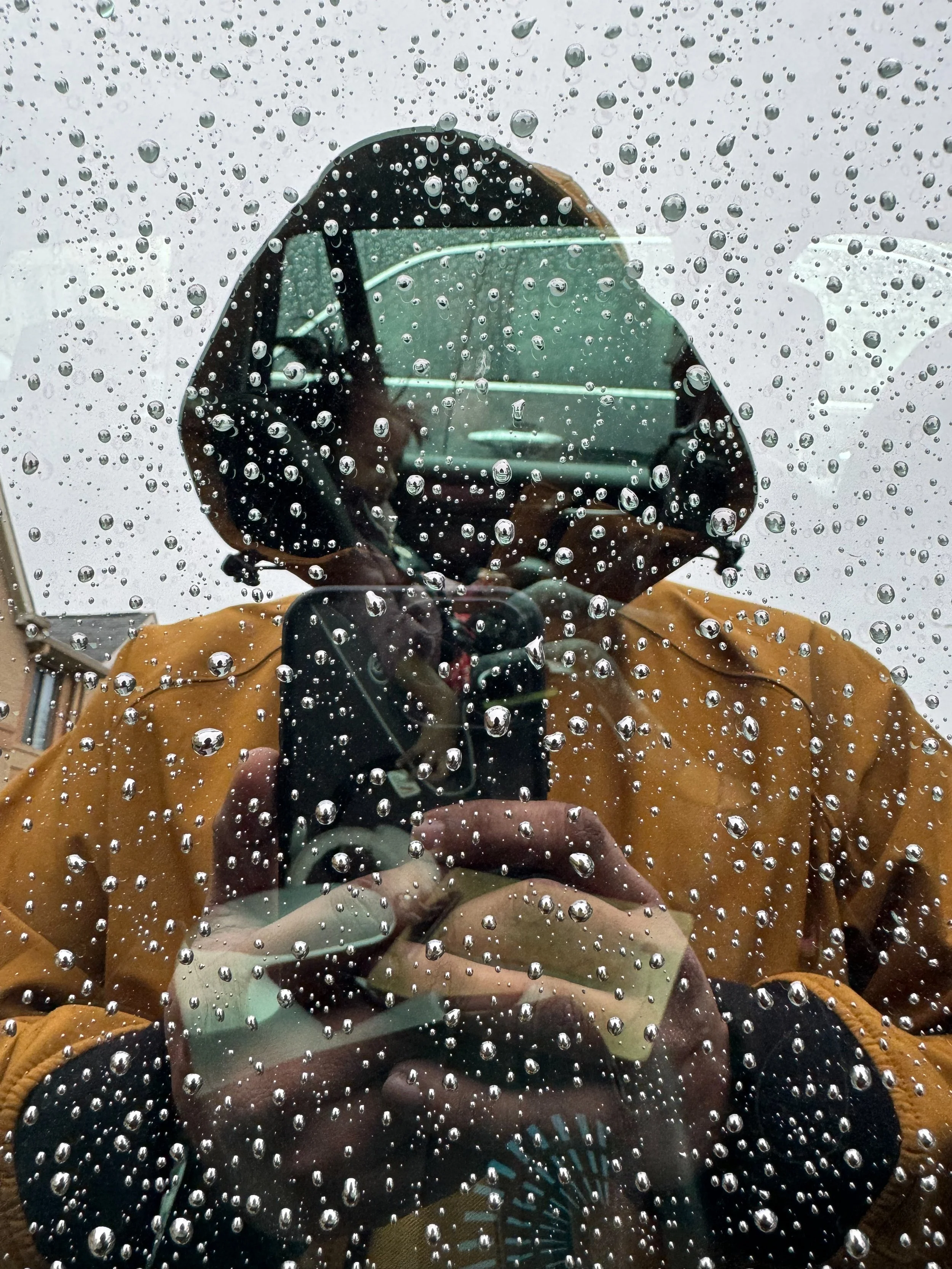 Photo of a person taking a picture through a rain-covered glass window, with a car and background scene reflected in the glass.