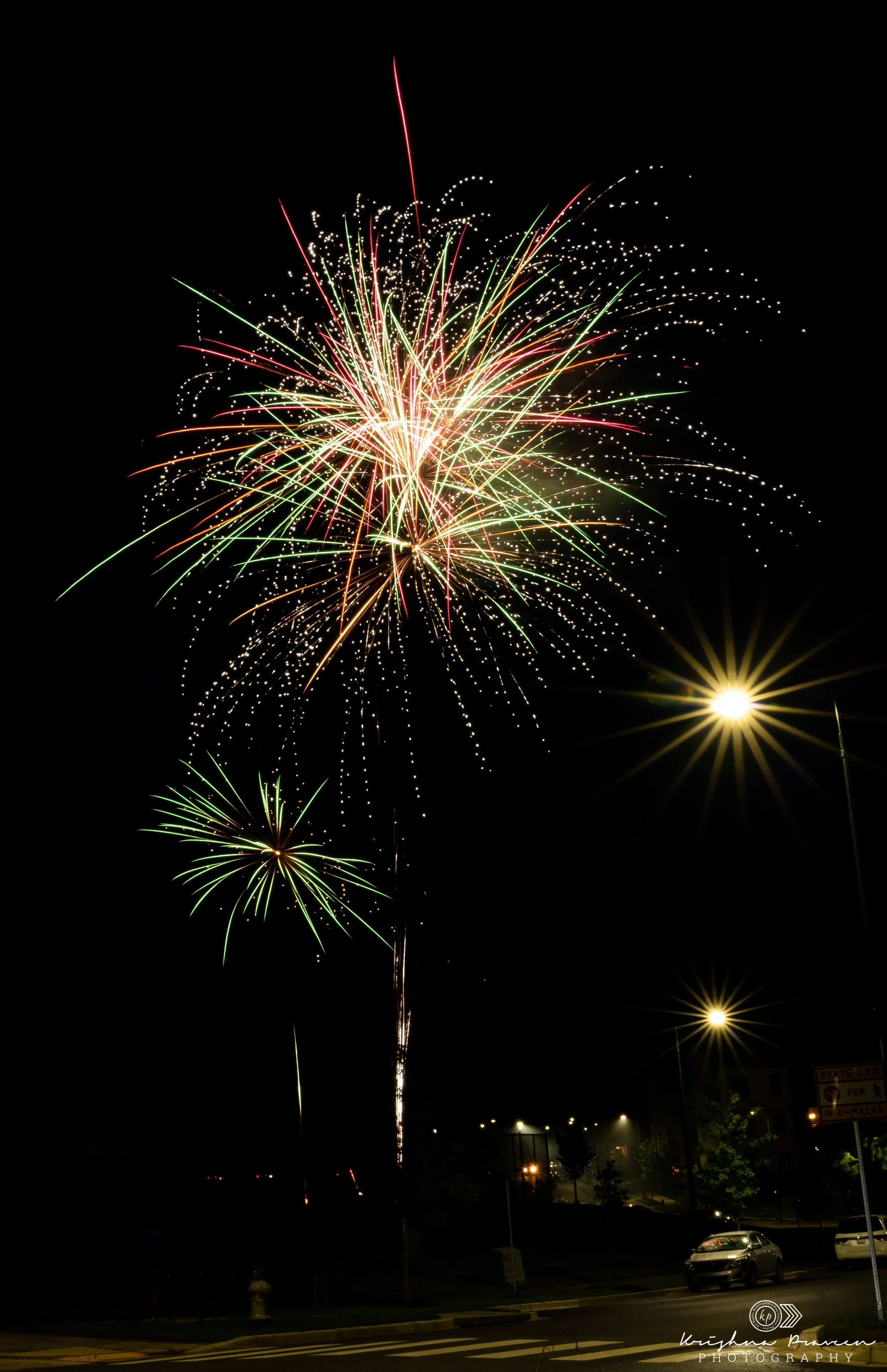 Colorful fireworks exploding in the night sky over a parking lot with cars and trees below.