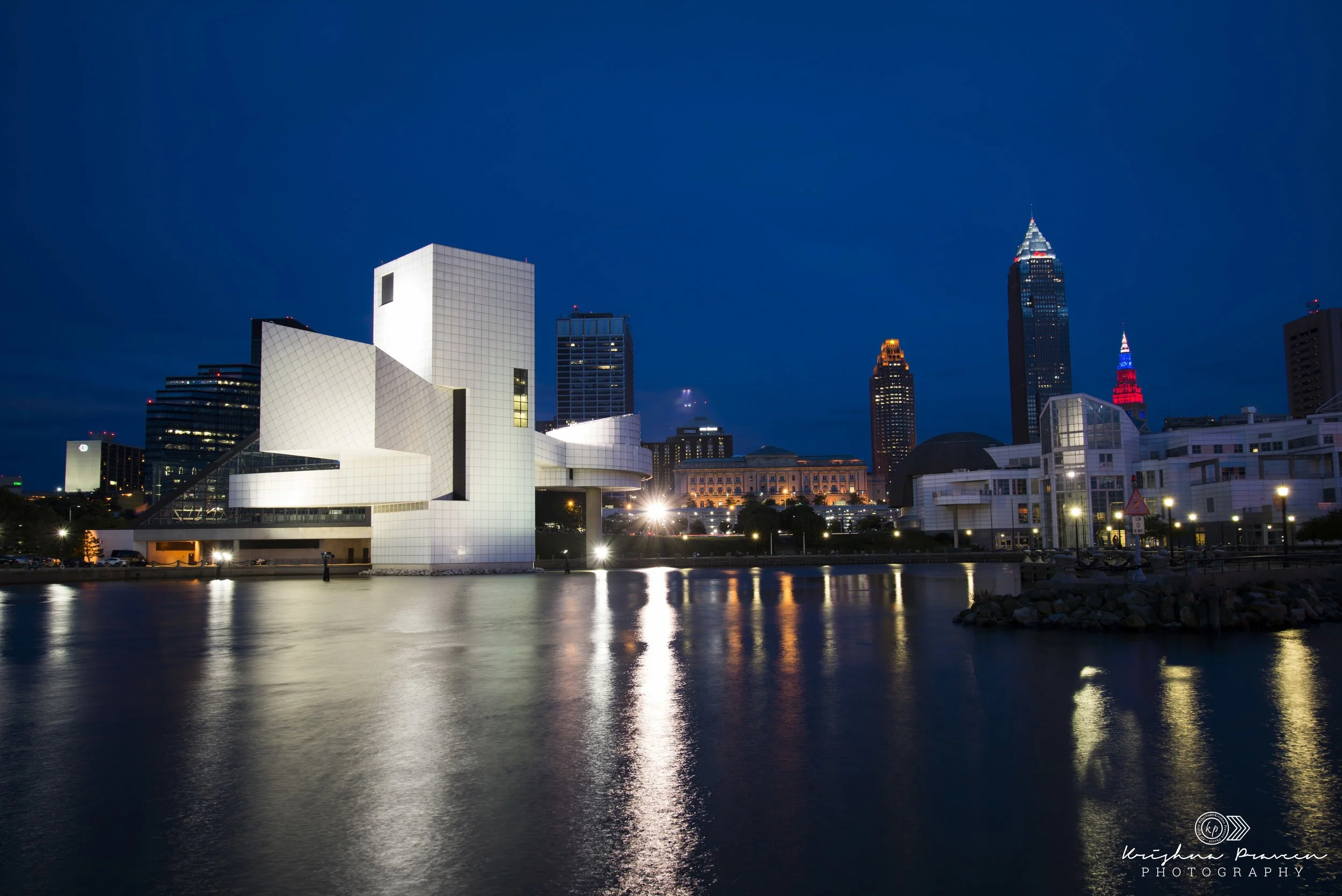 Nighttime cityscape of downtown with illuminated modern white building reflecting on river, historic buildings, and skyscrapers in the background.