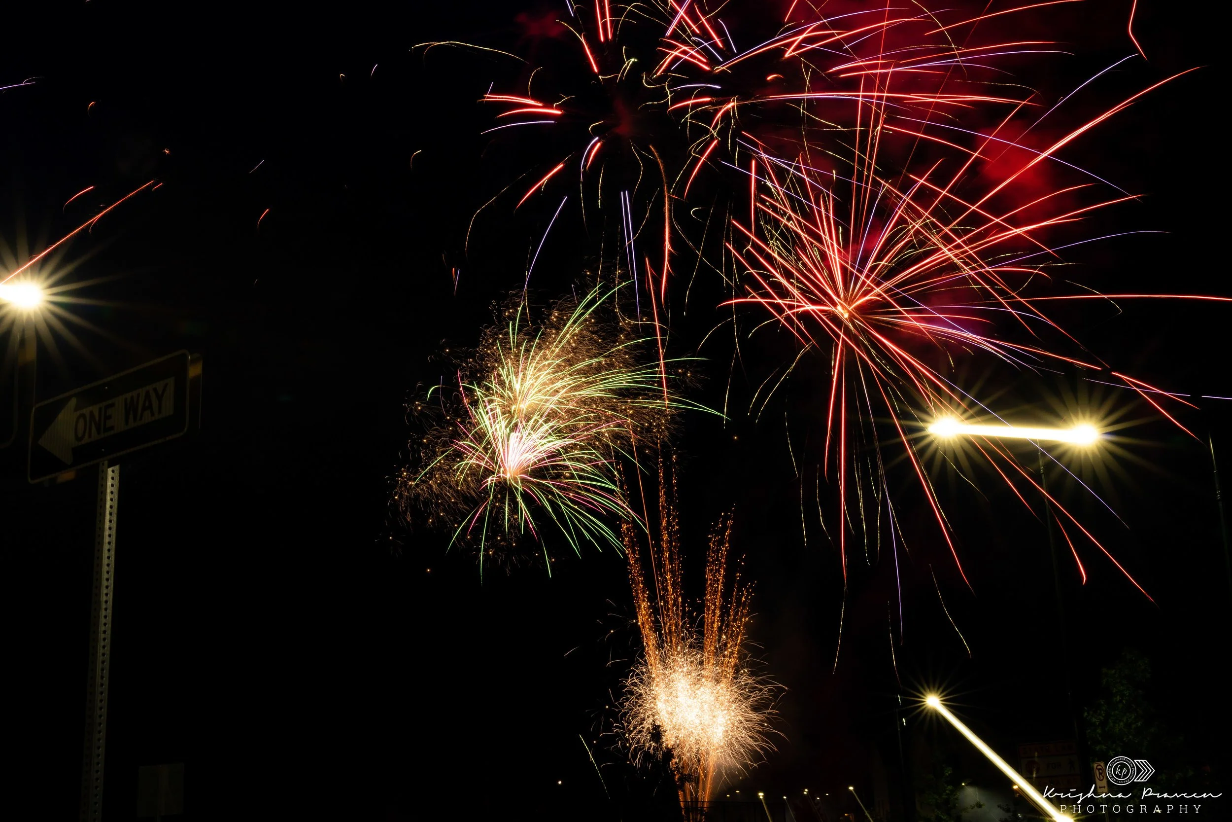 Fireworks exploding in the night sky with bright red, yellow, and purple streaks, and streetlights visible at the bottom.