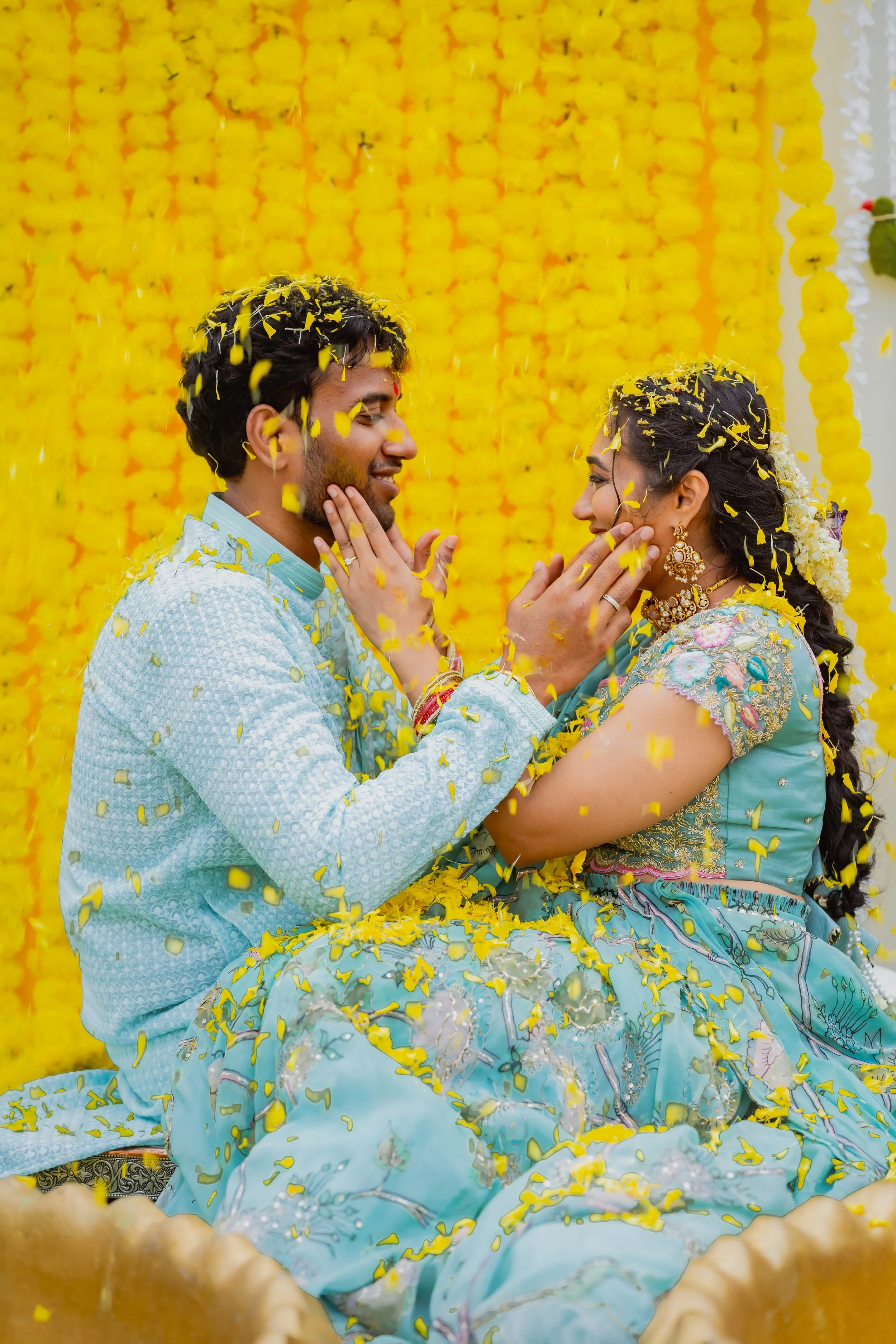 A man and woman, dressed in traditional colorful Indian clothing, are sitting together celebrating a wedding. They are smiling and touching each other's faces with yellow flower petals falling around them, against a backdrop decorated with yellow flo