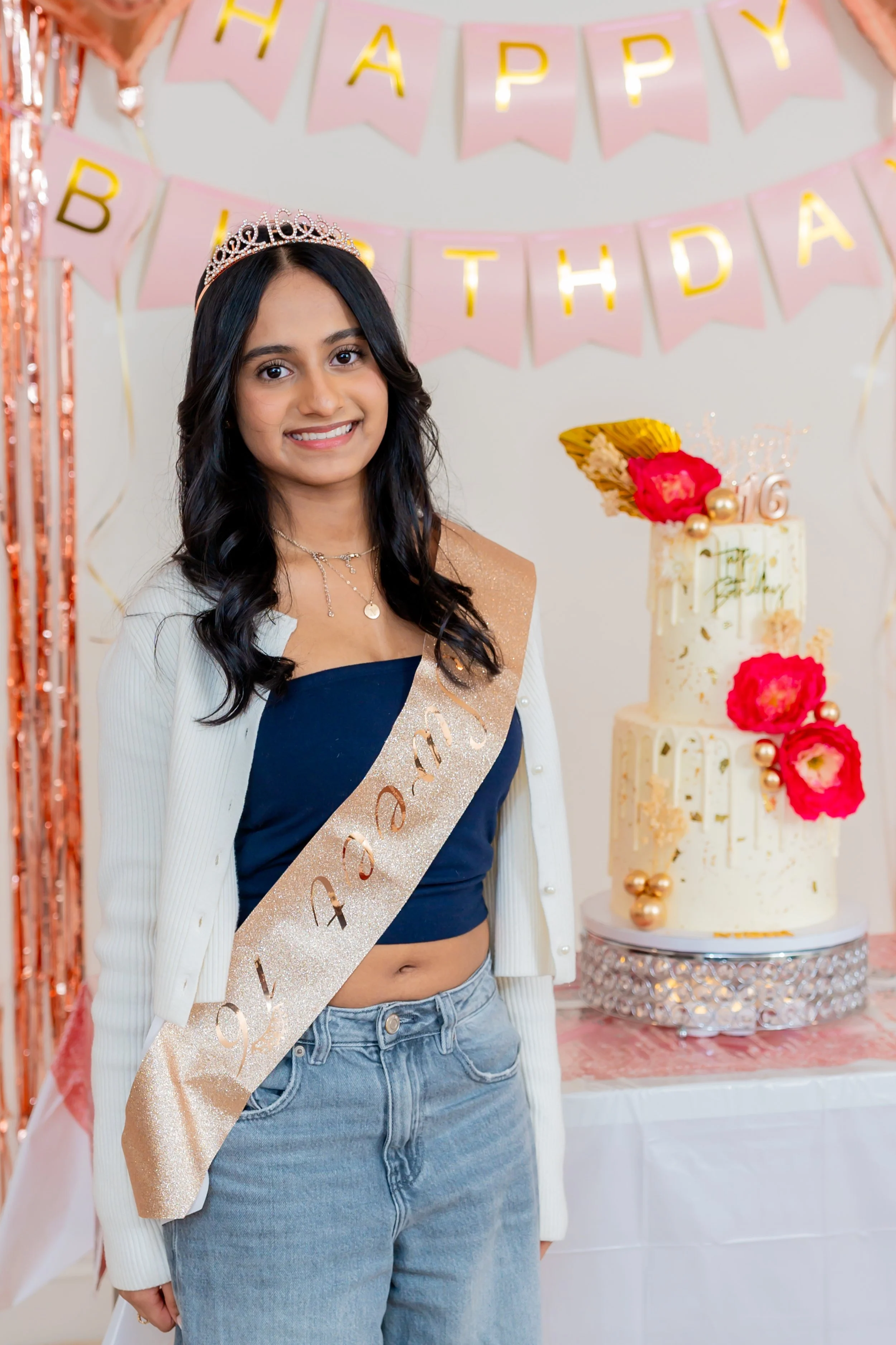 A young woman with long dark hair, wearing a tiara, a beige sash, a dark blue crop top, a white cardigan, and blue jeans, stands in front of a birthday party backdrop with pink and gold decorations, a cake, and envelope-shaped banners that spell out 