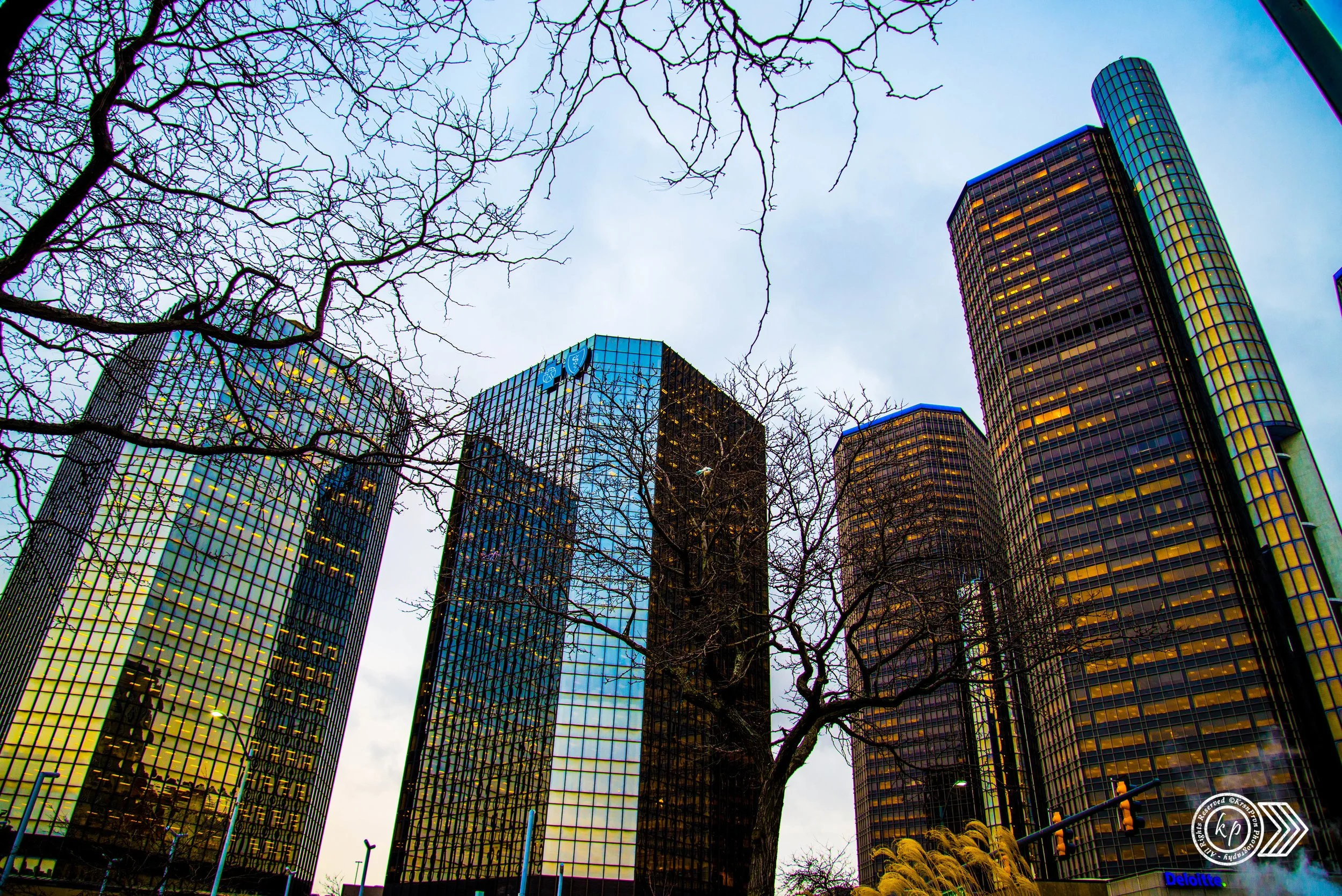 Tall modern glass skyscrapers reflecting the sky and city lights, with leafless trees in the foreground against an overcast sky.