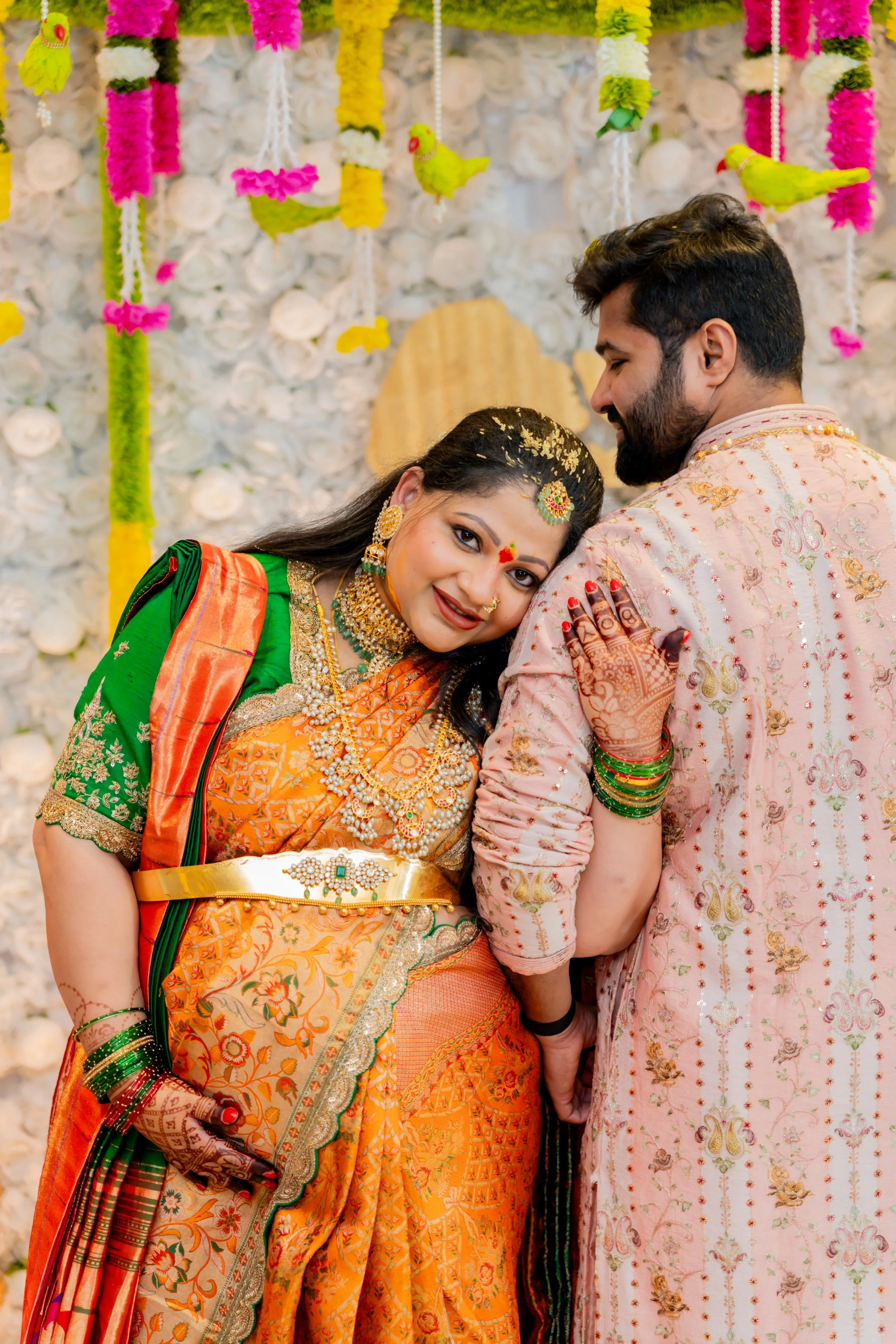 A bride and groom sharing a moment during their wedding celebration, dressed in colorful traditional Indian attire. The bride is wearing a green and orange saree with intricate jewelry, while the groom is in a cream-colored embroidered sherwani. The 