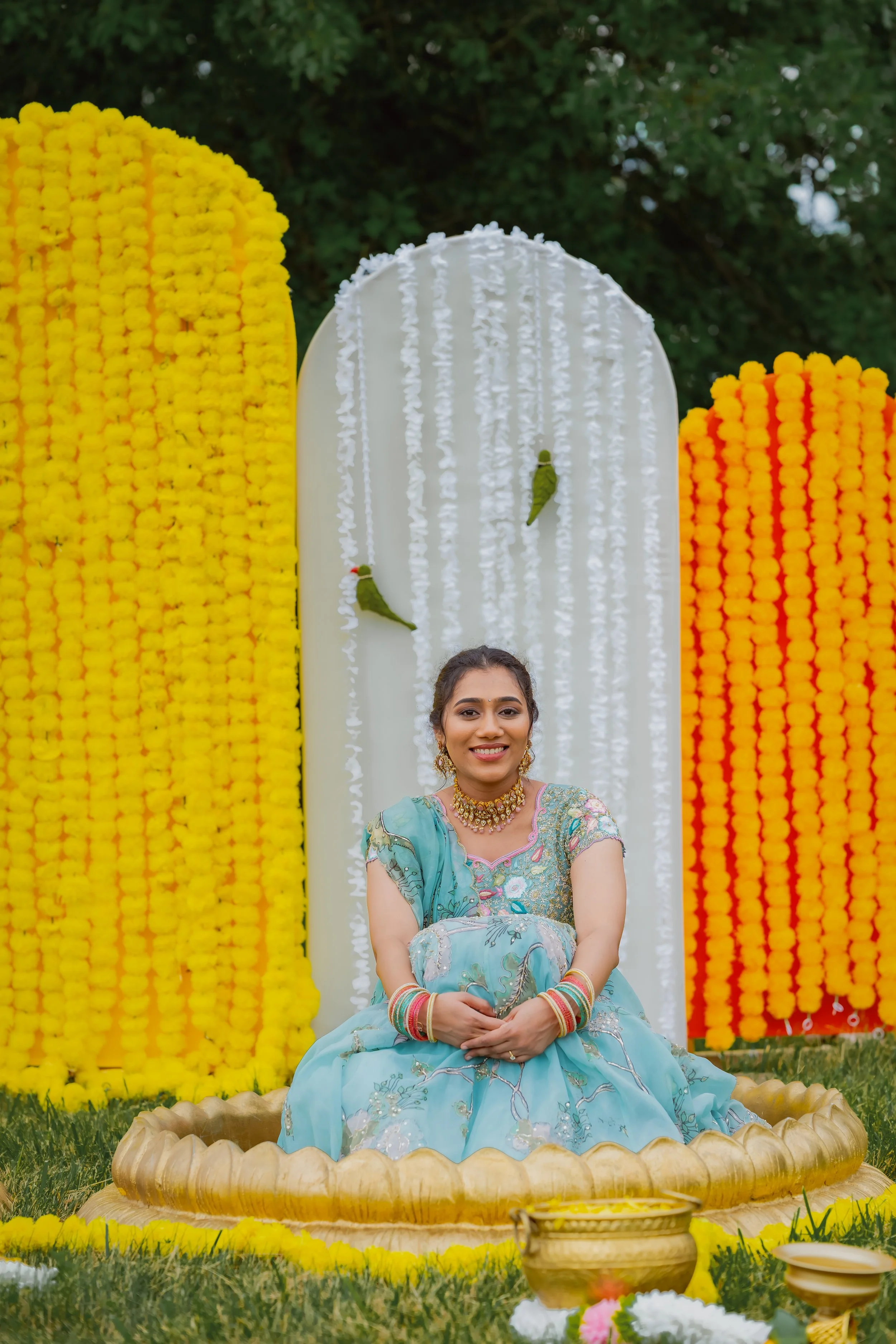 A young woman dressed in traditional South Asian attire, sitting on a decorative platform during a festive outdoor event decorated with yellow and white floral arrangements, with greenery and trees in the background.