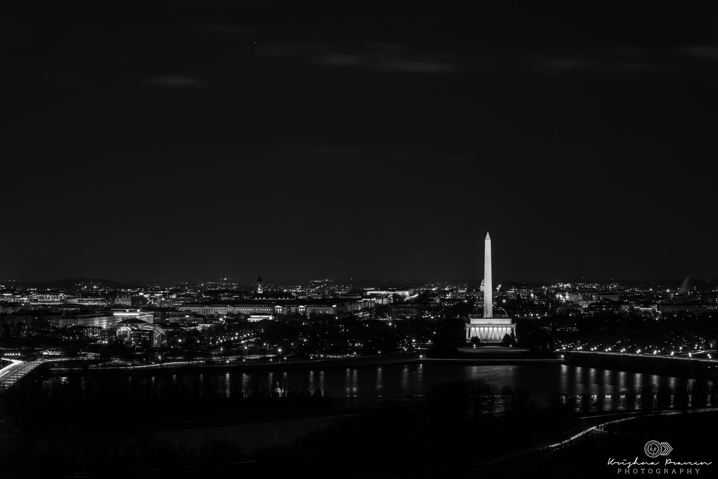 Nighttime city view of Washington, D.C., with the Washington Monument illuminated in the distance, dark sky with a few clouds, and city lights reflecting on the water.