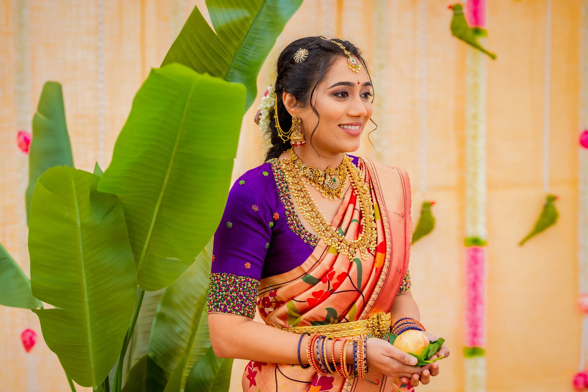 A woman dressed in traditional Indian attire and jewelry, smiling, surrounded by large green leaves and a decorated background.