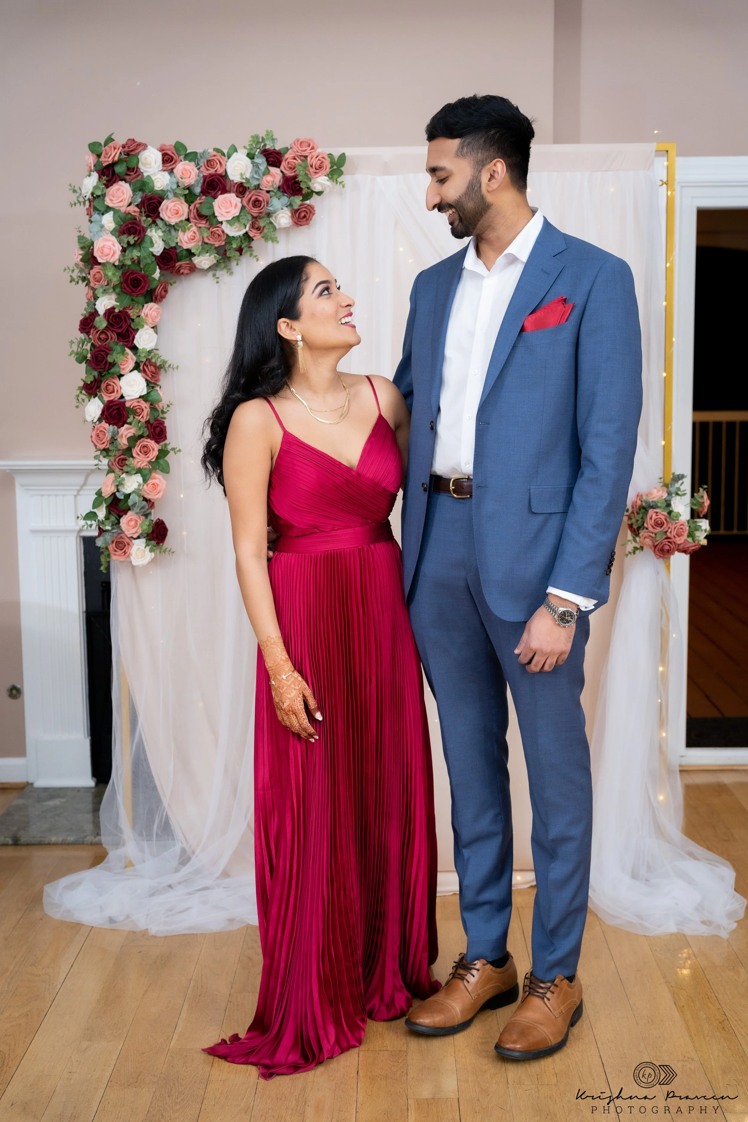A couple standing together at a celebration, with the woman in a long, red, pleated dress and the man in a blue suit with a red pocket square. They are smiling at each other in front of a floral backdrop decorated with pink, red, white roses, and gre