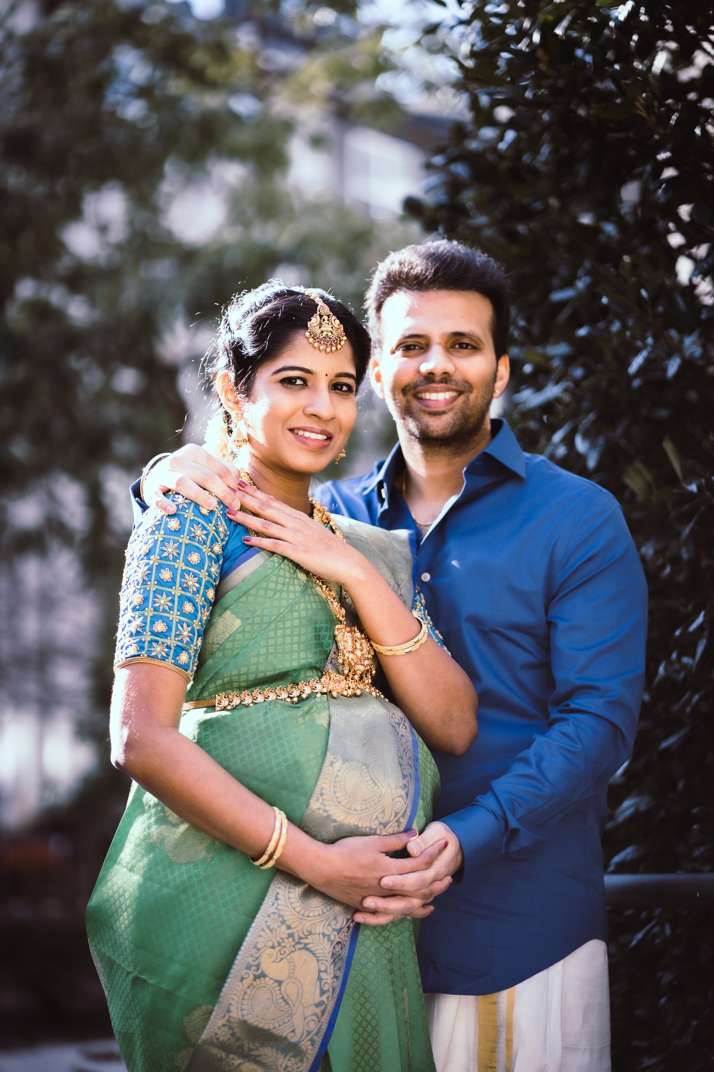 A smiling couple, dressed in traditional Indian attire, stands outdoors with lush greenery in the background. The woman wears a green saree with gold and blue accents, jewelry, and has her hand on her partner's shoulder. The man is dressed in a blue 