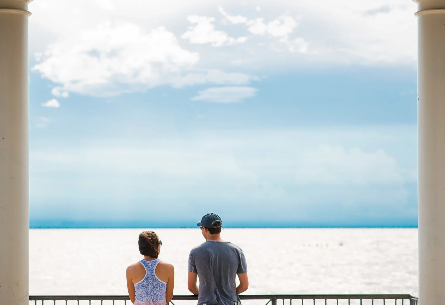 A man and a woman stand side by side on a balcony overlooking a beach, with the ocean and a cloudy sky in the background.