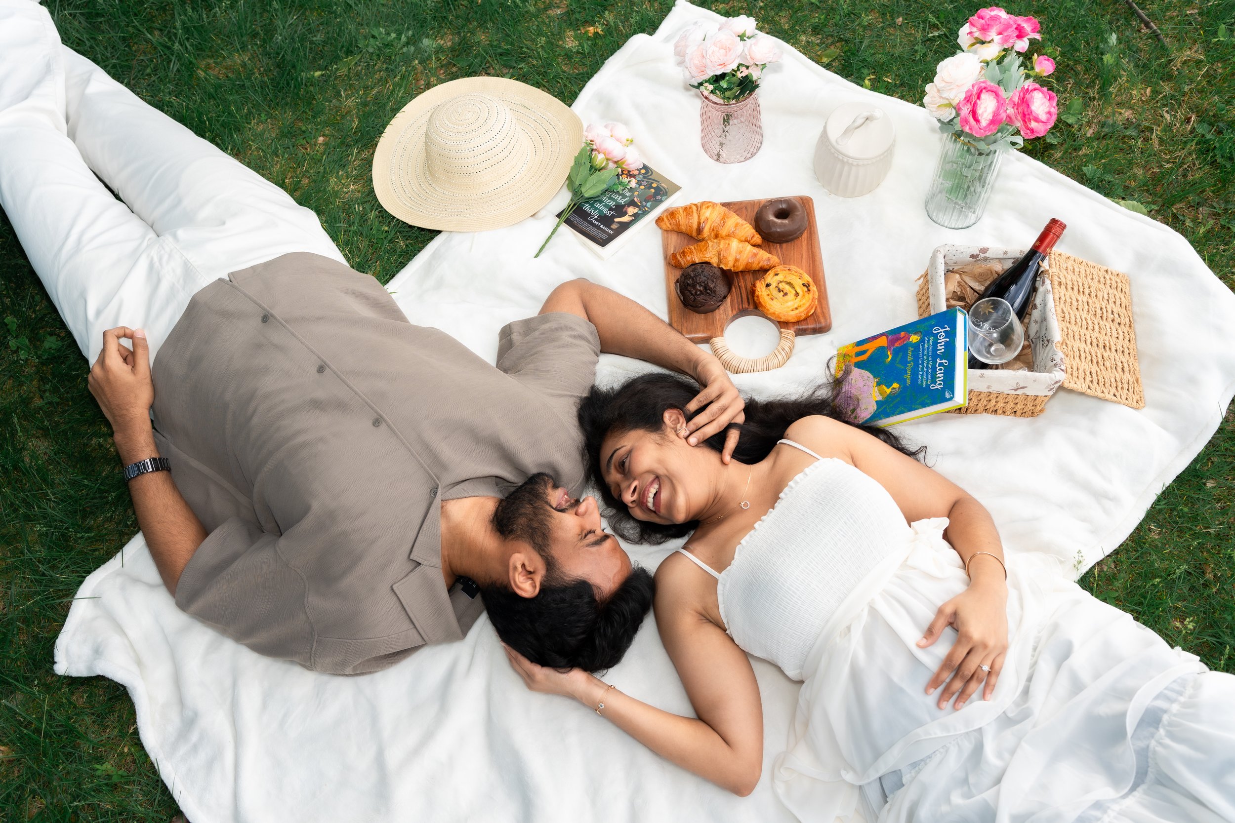 A man and woman lying on a white blanket outdoors, smiling and looking at each other. The scene includes vases with pink flowers, a book, pastries, a bottle of wine, and a glass. It appears to be a picnic on grass.