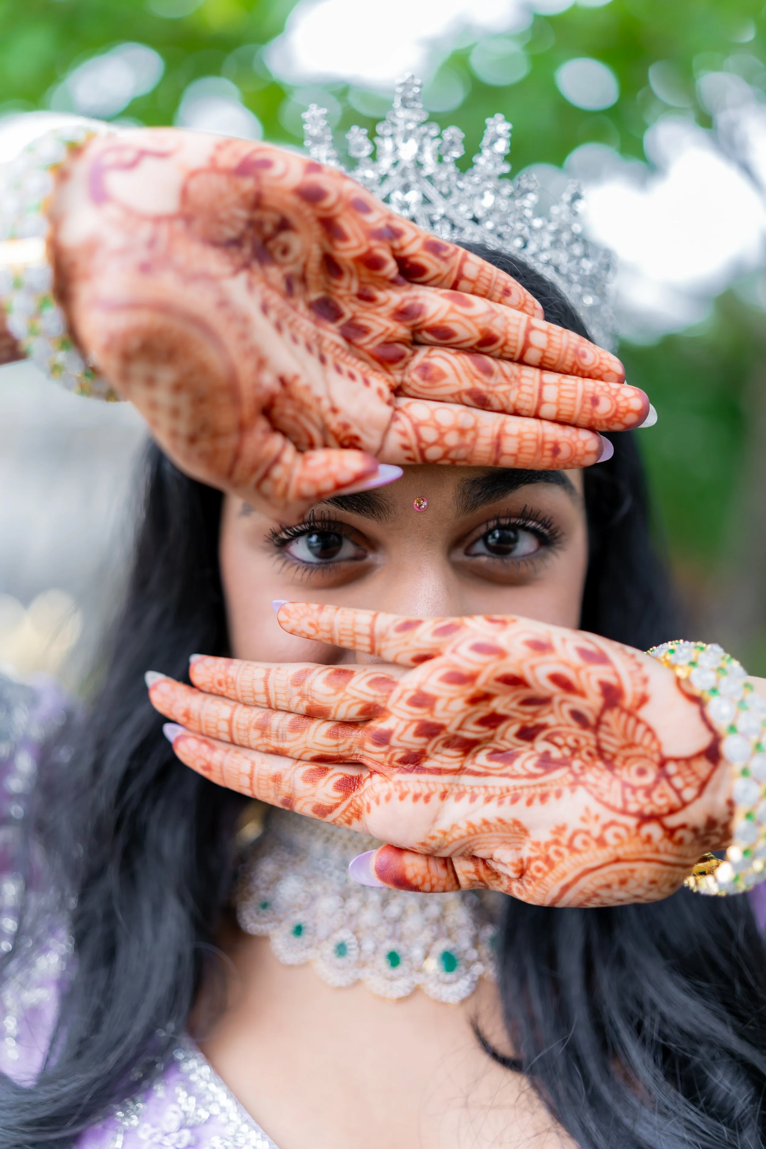 A young woman with dark hair, wearing a silver tiara, has henna designs on her hands which she is holding up in front of her face. She is also wearing a jeweled choker necklace and a bindi on her forehead, with a blurred green background.