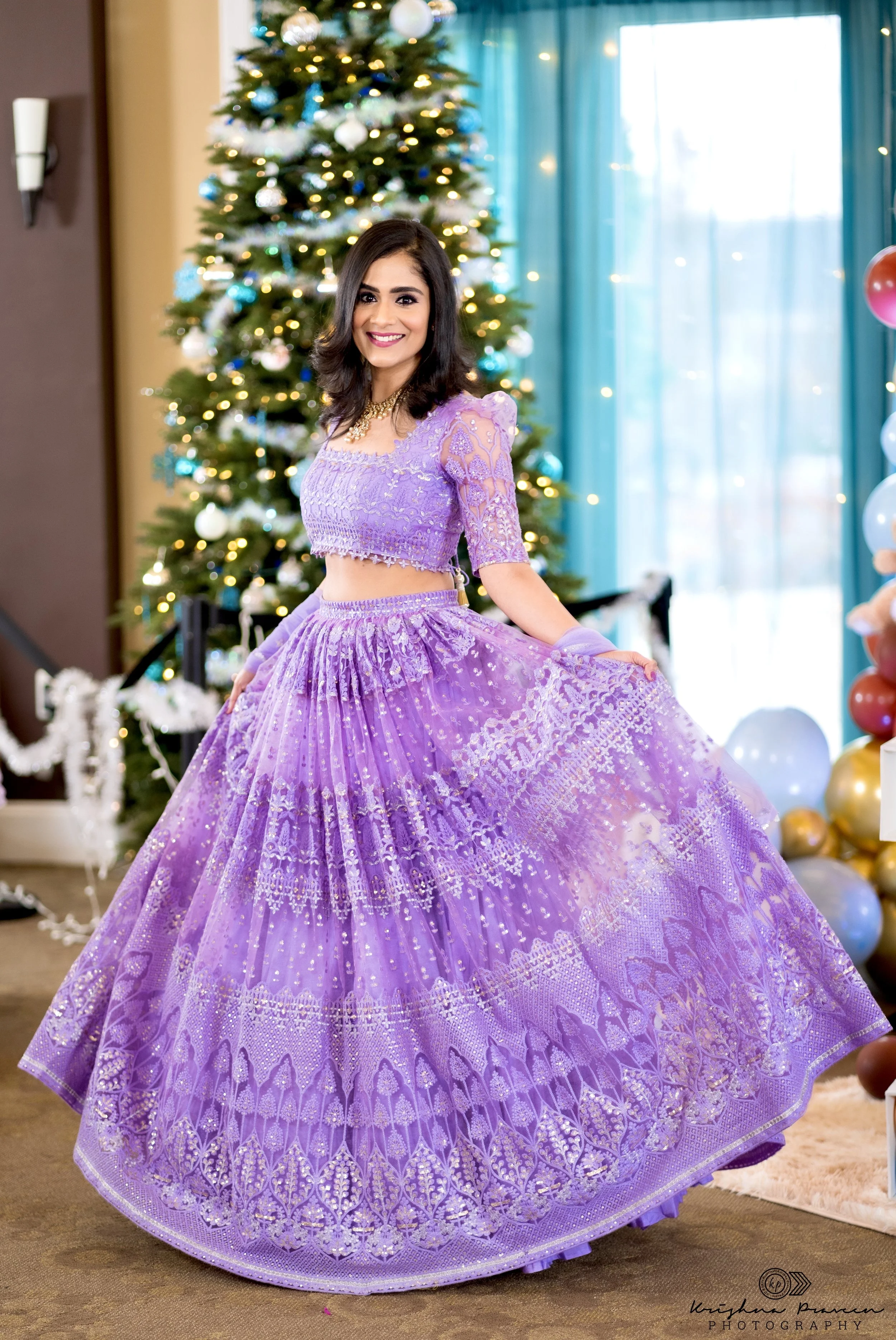 A woman dressed in a purple, intricately embroidered lehenga, standing indoors in front of a Christmas tree decorated with white, blue, and silver ornaments and lights. She is smiling and has dark, shoulder-length hair.