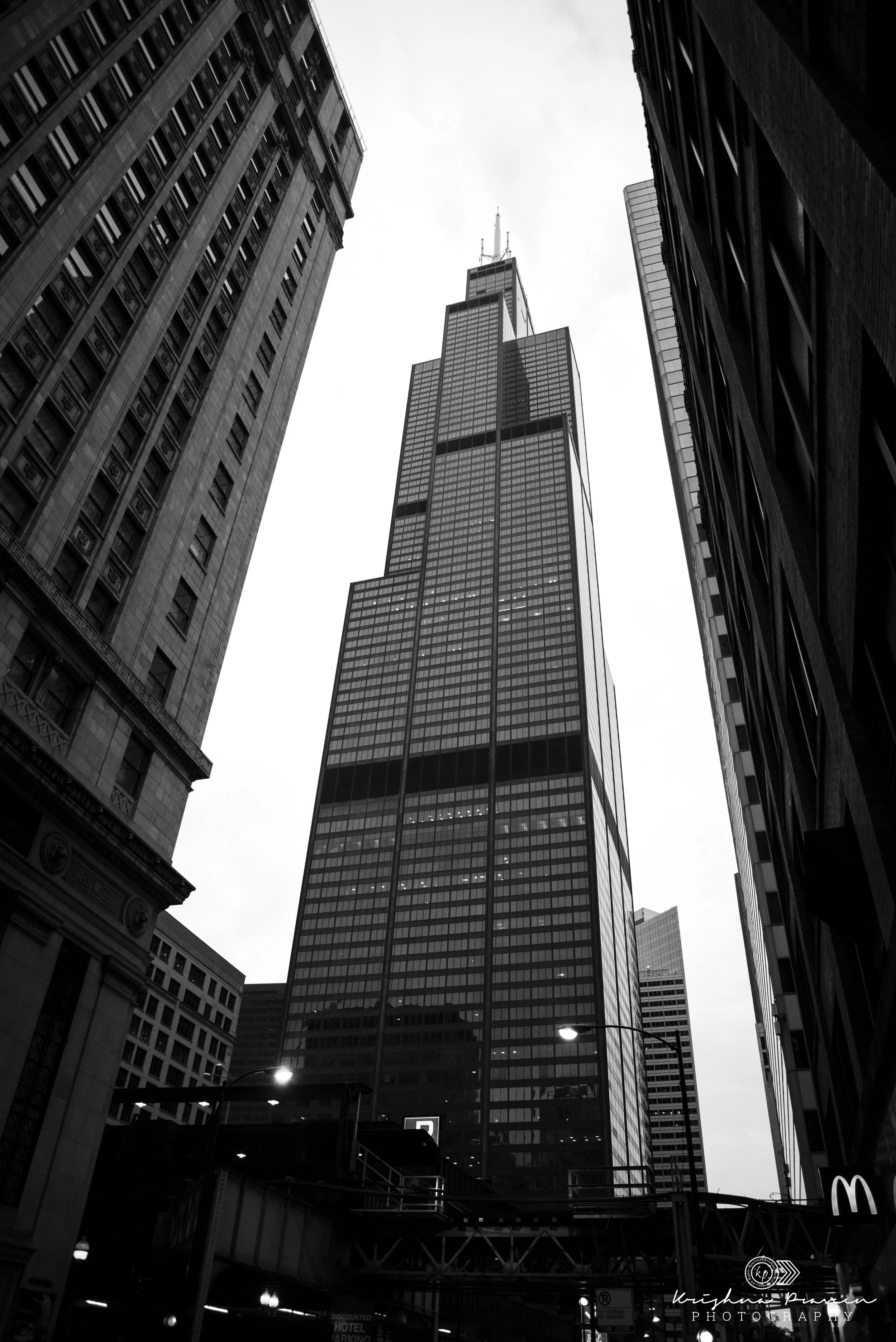 A black and white cityscape view looking up at tall skyscrapers in downtown. The image features a prominent modern building with glass windows, flanked by older buildings on either side. The sky above is overcast, and streetlights are visible at the 