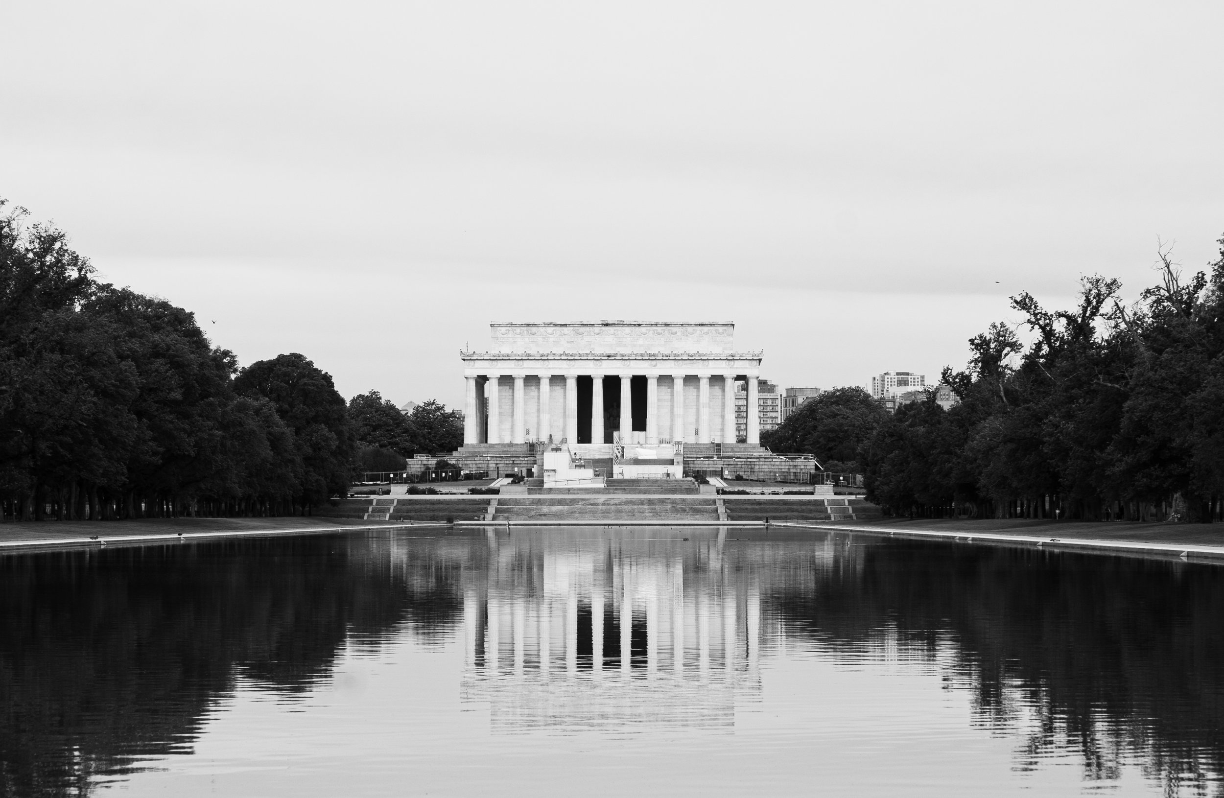 A black-and-white photo of the Lincoln Memorial in Washington, D.C., with trees on either side and reflections in the water in the foreground.
