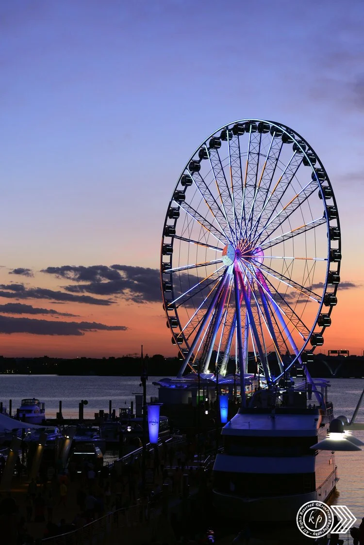 A large illuminated Ferris wheel by the water at sunset, with boats docked nearby and a colorful sky with clouds at dusk.
